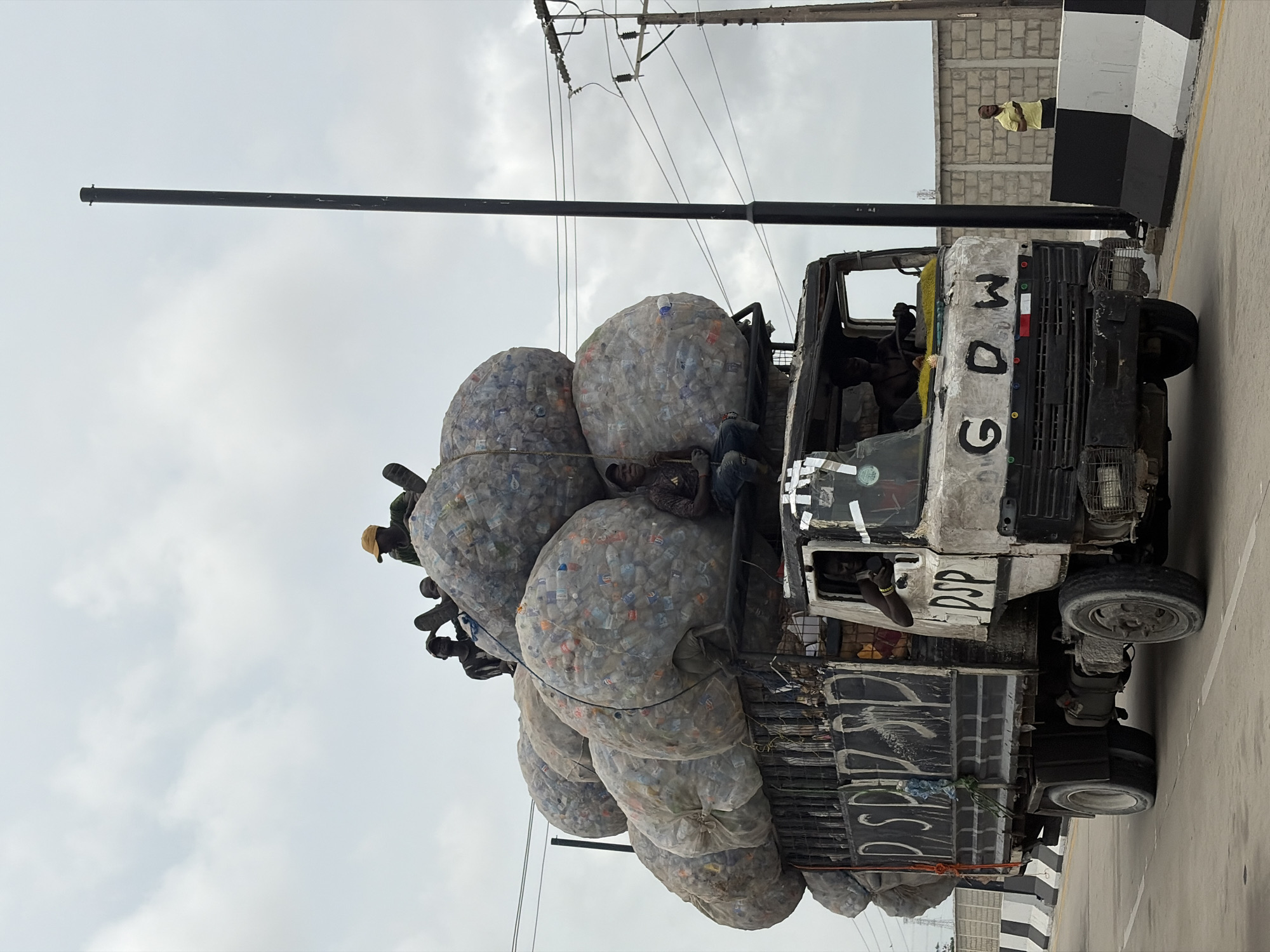 A heavily overloaded truck with several large bundles of compacted plastic bottles is on the side of a road, likely in Igboefon, Nigeria. Several people are riding on top of the load of plastic bottles. The truck is heavily graffitied. Another individual wearing a yellow shirt is standing on the side of the road. Overhead utility lines and a cloudy sky are visible.