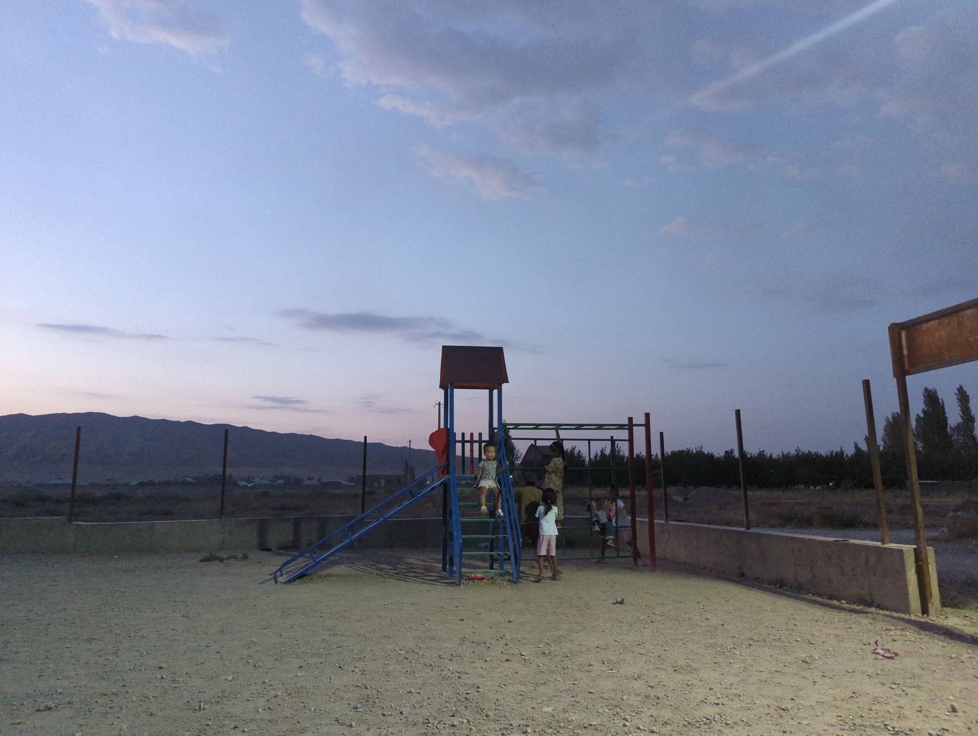 In the city of Üch-Döbö, Kyrgyzstan, at dusk, an outdoor playground features a blue metal slide and climbing structure. Several children are present. One child, wearing a light-colored shirt and shorts, is on the slide. Another child in a white shirt and pink shorts is standing near the base of the structure. A third child, wearing a white shirt and shorts, is climbing the monkey bars. A fourth child in a striped shirt is also on the monkey bars. An adult in a patterned dress is standing near the monkey bars, appearing to supervise. The playground is situated on a sandy surface, enclosed by a low concrete wall and a chain-link fence. In the background, mountains are visible under a pale blue, cloudy sky. The scene suggests a casual, everyday moment of children playing outdoors as the day ends.
