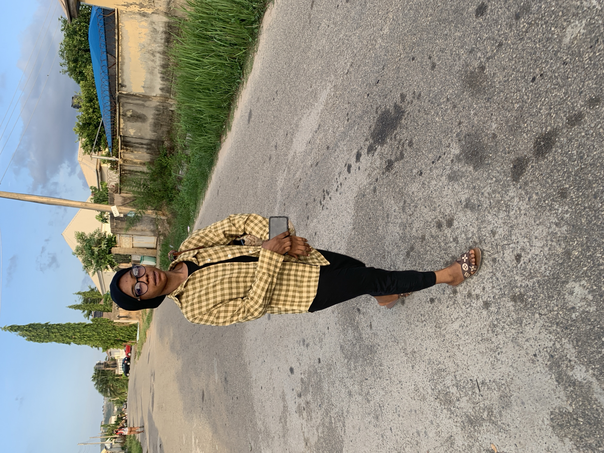 A young woman stands on an asphalt road, holding a mobile phone in her hands. She wears black trousers, a black top, a yellow and brown checkered jacket, a black beanie, and glasses. She also wears flat sandals with a brown and flower pattern.
The environment consists of a residential area with buildings, trees, and vegetation along the roadside. There is a building with a blue awning in the background. The road surface is cracked and shows signs of wear. The sky is visible in the distance.
The location is most likely a residential area in Abuja, Nigeria. The photograph shows a casual, informal scene.