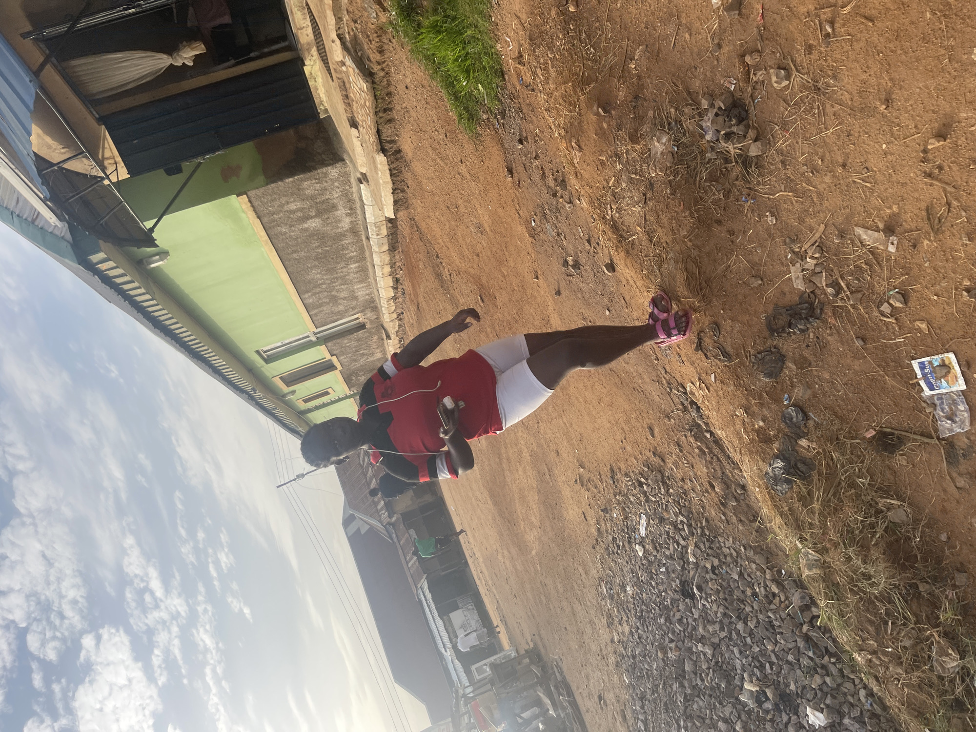 A medium shot captures a young Black woman walking on a dirt path. She is wearing a red and black striped t-shirt, white shorts, and pink sandals. She is holding a phone in her right hand and has earbuds in her ears. The woman is walking towards the camera, and her body is angled to the right.
The background shows a street lined with buildings. The building directly behind the woman is painted green and has a corrugated metal roof. Other buildings in the background appear to be made of concrete and corrugated metal. There are other people walking in the background, though they are out of focus. The ground is a mix of dirt, gravel, and dry grass. The sky is partly cloudy.
The image appears to have been taken in a rural or semi-urban area of Rafin Par, Nigeria, given the style of buildings and the unpaved road. The overall impression is of a casual outdoor scene on a bright, possibly warm day.