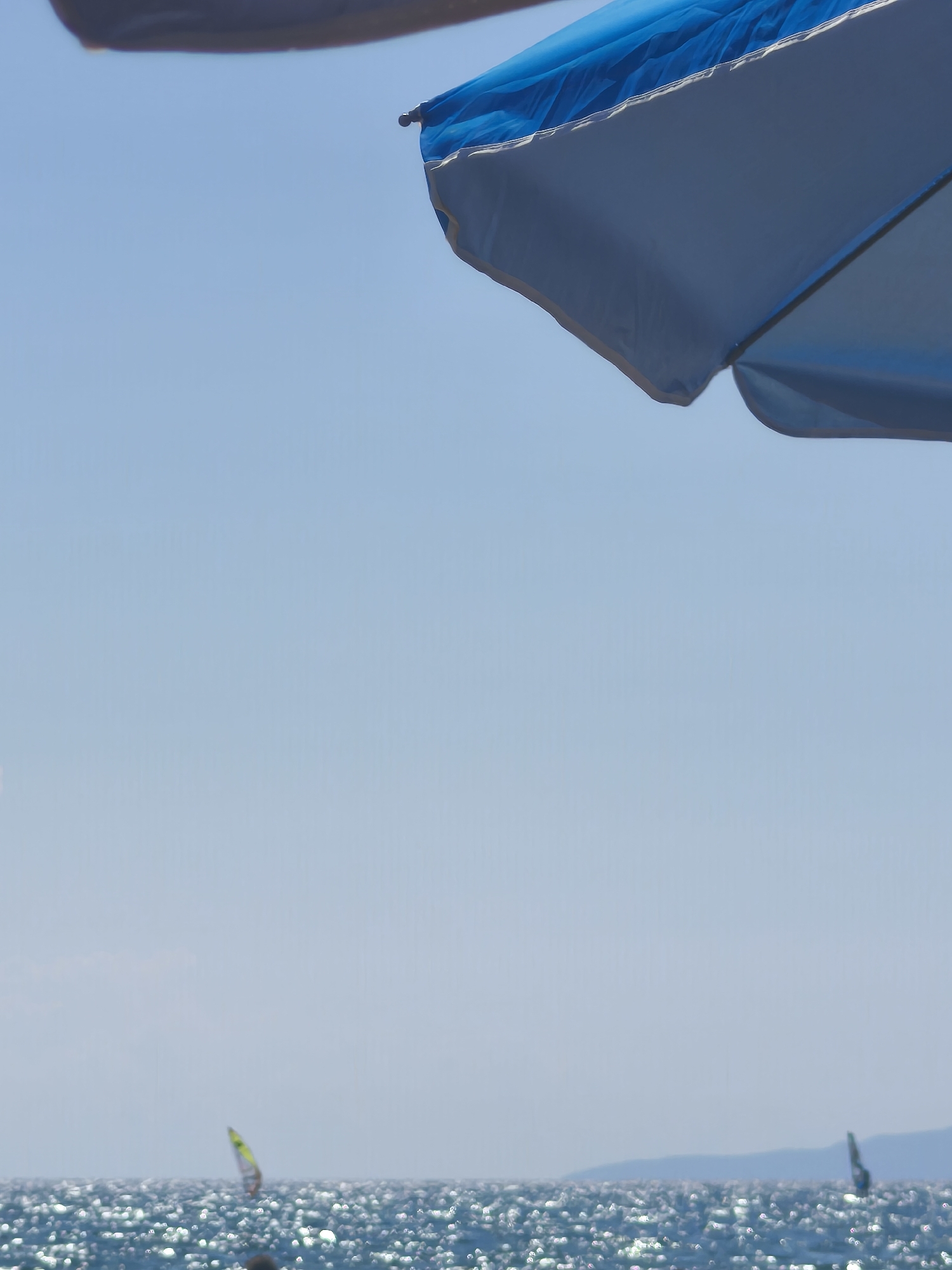 The image shows a view of the sea from a beach in Castiglione della Pescaia, Italy. A blue and white beach umbrella occupies the top portion of the frame, casting a shadow. The sea is visible in the lower part of the image, with sunlight glinting off the surface. Two windsurfers are in the distance on the water. The sky is clear and blue.