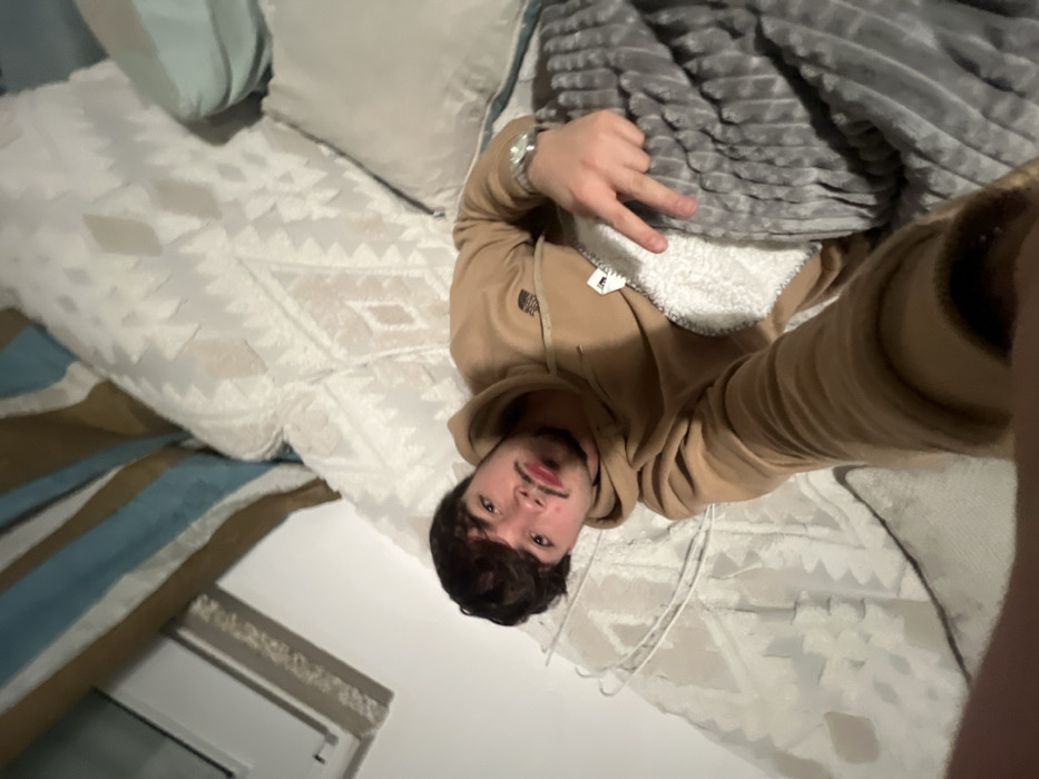A young man wearing a tan hooded sweatshirt lies on a patterned white and beige comforter. A gray blanket is partially covering him. He holds up two fingers in a peace sign gesture. His face is in focus, showing dark hair, a mustache, and fair skin. Pillows are in the background, and a window with a white frame is visible in the lower-left corner. The image is taken from a first-person perspective, slightly angled.