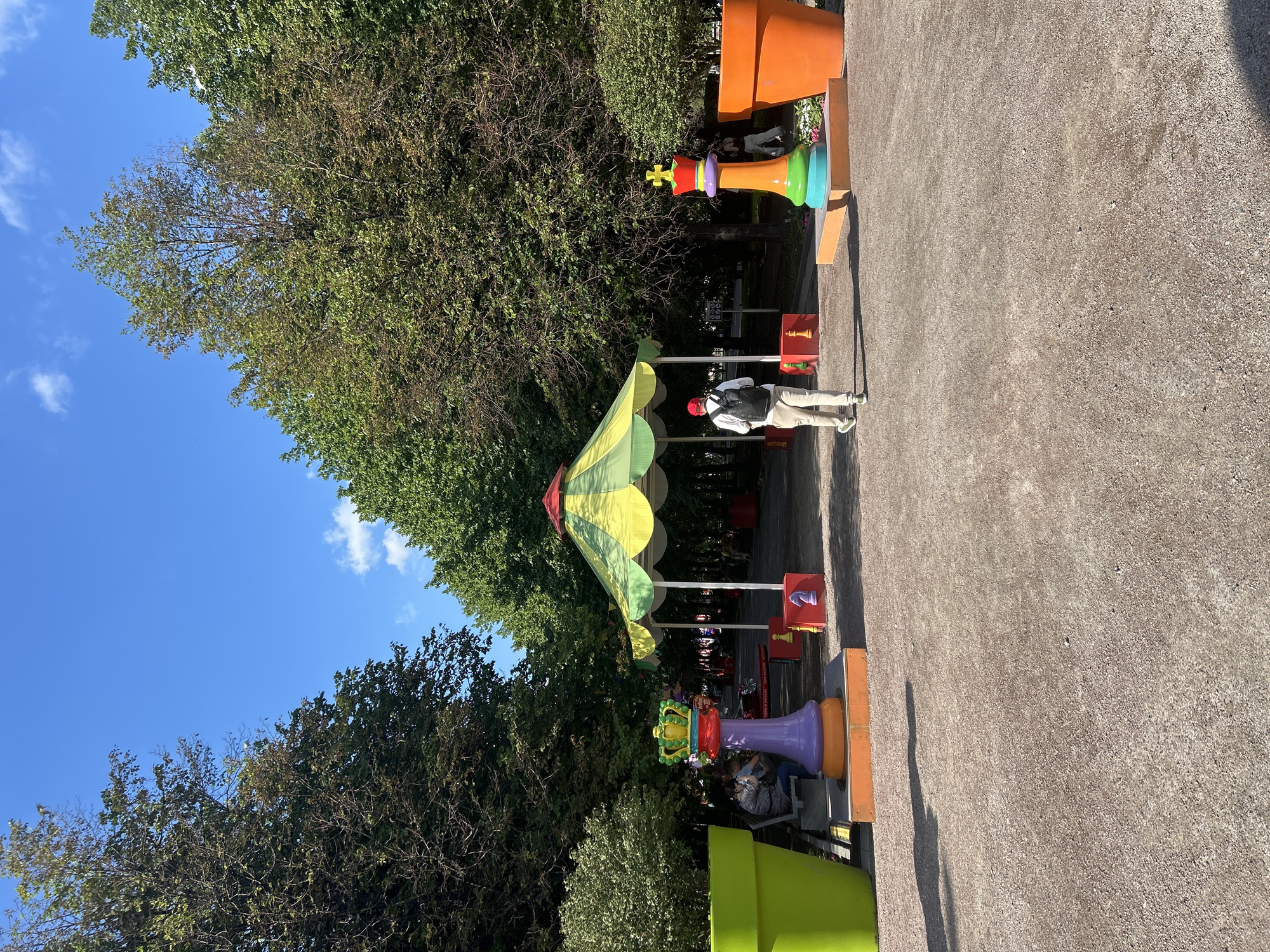 An outdoor park scene with a pathway lined by large trees on both sides. In the foreground, a person wearing a red cap and a backpack walks away from the camera down a gravel path. To the left and right of the path are two oversized, brightly colored king chess pieces. Behind the chess pieces are enormous, colorful planters, one lime green and one orange. In the middle ground, a yellow and green striped canopy tent is set up over a seating area. There are several people seated on benches under the tent. Several other oversized chess pieces are visible around the tent. The sky is blue with a few scattered white clouds.