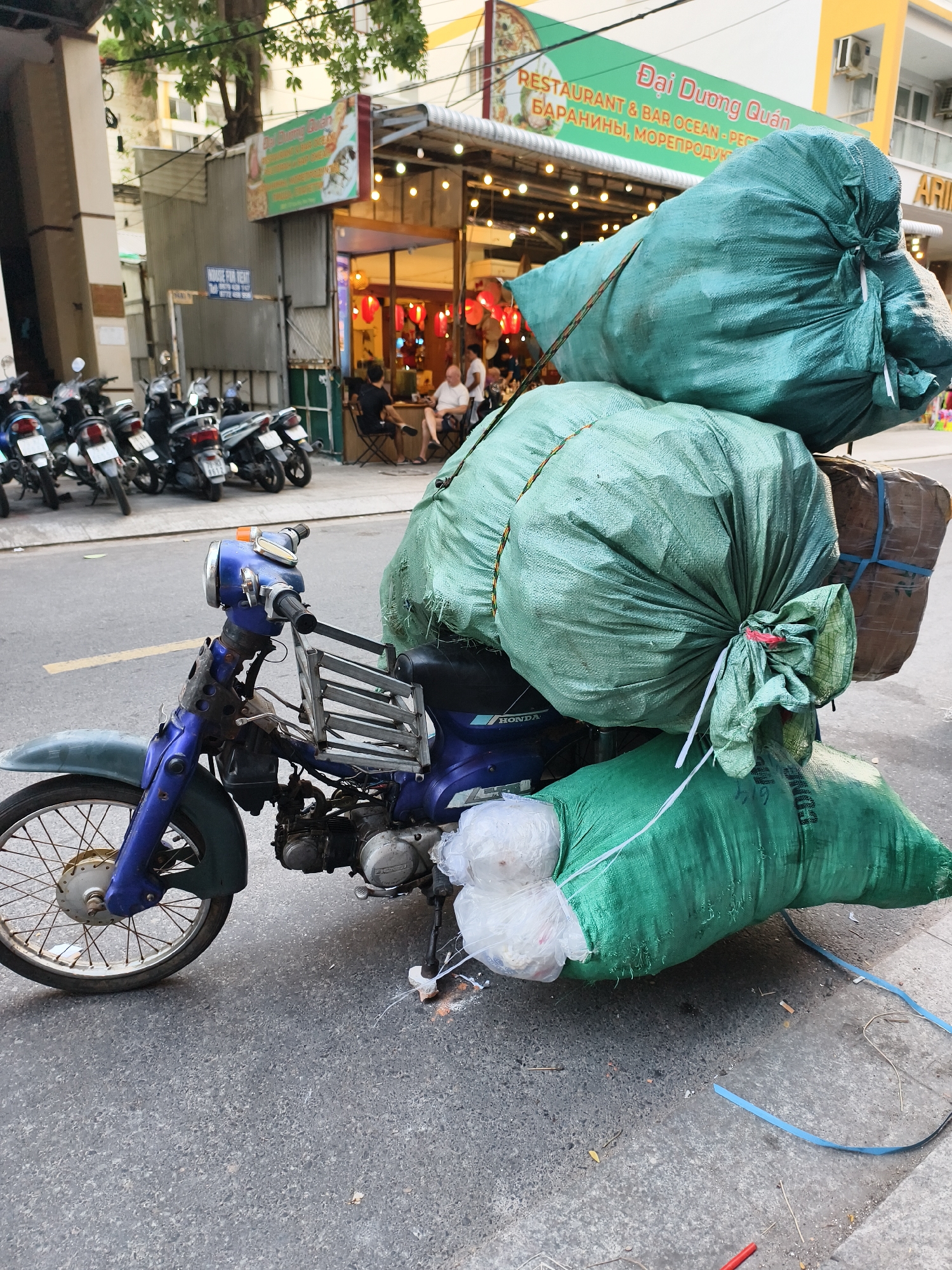 The image captures a blue Honda motorcycle heavily laden with four large, tightly packed, green sacks. Two of the sacks are secured on the seat, one behind the seat, and another at the front near the handlebars. The sacks appear to be filled with light-colored material, possibly plastic.
The motorcycle is parked on a paved street in what appears to be a city in Vietnam, as indicated by the signage on a nearby restaurant, "Đại Dương Quán RESTAURANT & BAR OCEAN." The restaurant has outdoor seating with people sitting at tables under hanging lanterns. Several parked motorcycles are lined up on the street further away from the camera. The environment is urban, featuring buildings and overhead wires. The sky is overcast.