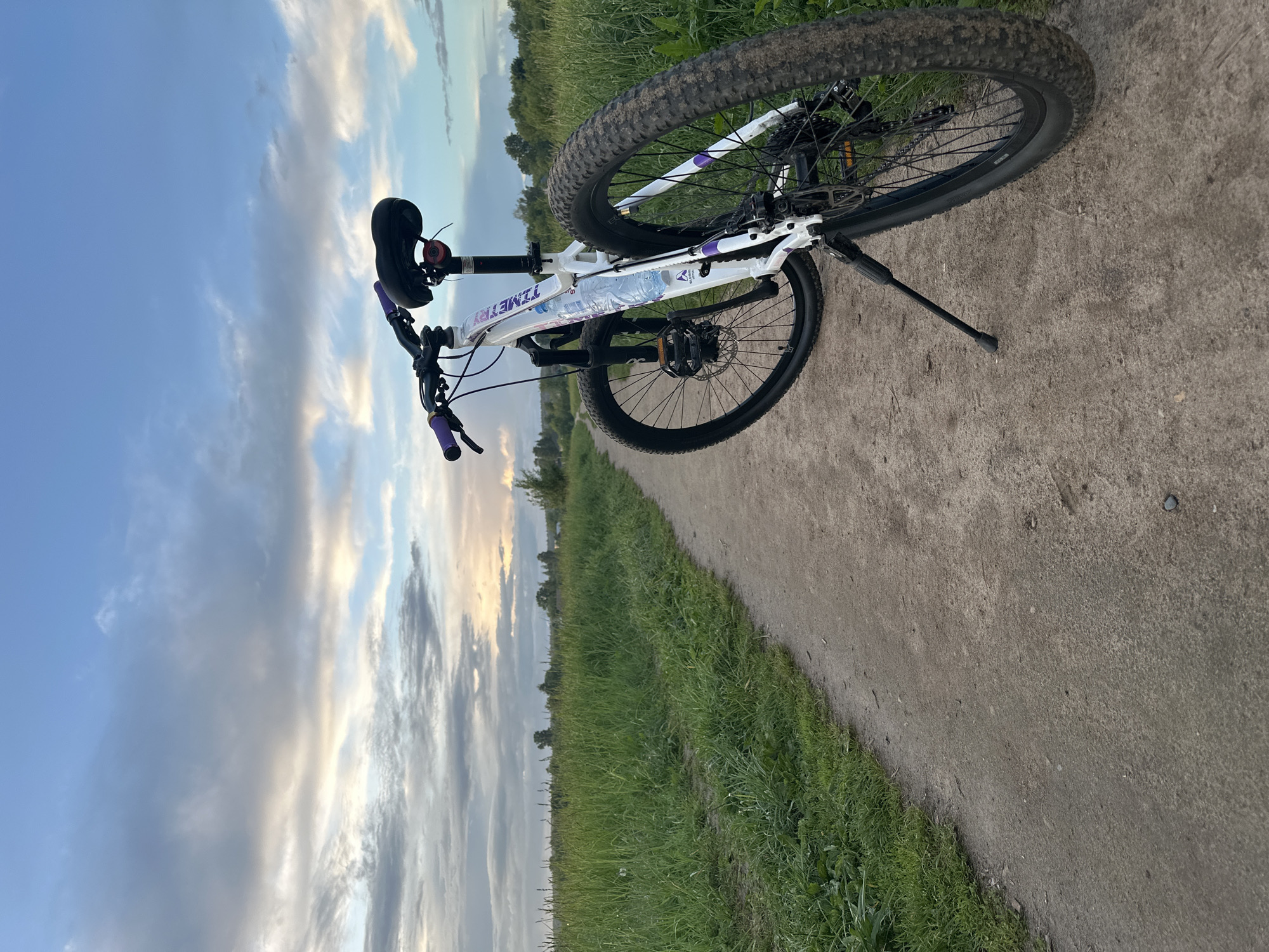 A white mountain bike with purple accents is parked on a dirt path next to a grassy field. The bike has a kickstand deployed, and a water bottle is visible in its frame. The path is made of packed dirt or gravel. The grassy field extends towards the horizon, with trees in the background. The sky is visible in the upper left of the frame, displaying shades of blue and light clouds. The image appears to be taken at dusk or dawn due to the lighting. The bike brand is "Timetry." Based on the location cues, the image might be taken near Shchyolkovo, Russia.