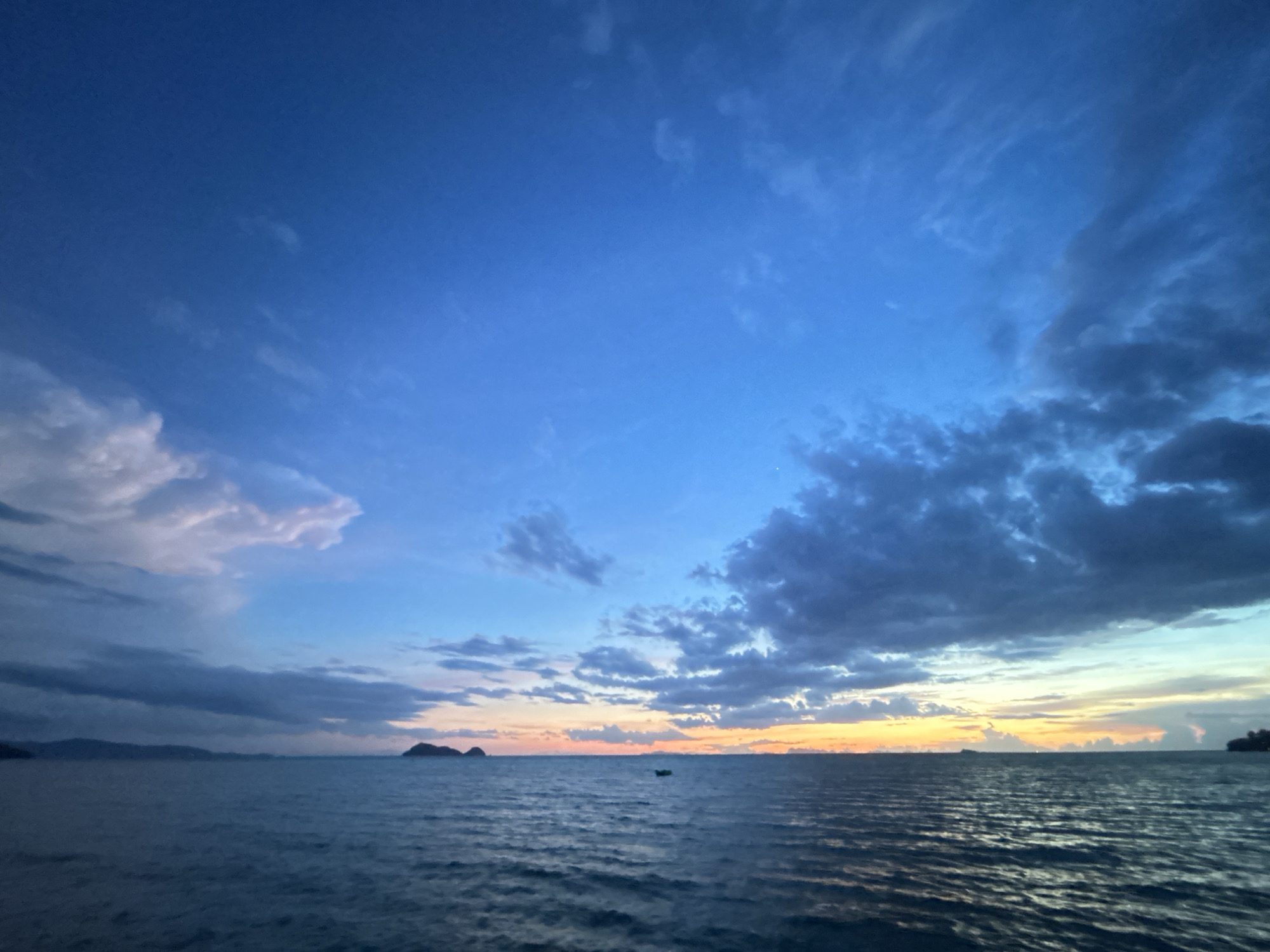 The image depicts a tranquil seascape at dusk, likely captured from the shores of Ko Pha-ngan District, Thailand. The sky is a dramatic gradient of deep blue, transitioning to lighter blues and hints of orange and yellow near the horizon, suggesting the sun has recently set or is about to rise. Scattered clouds, some dark and heavy, others wispy and light, add texture and depth to the celestial canvas.
The sea itself is a deep, calming blue, with gentle ripples reflecting the colors of the sky. A small, dark silhouette of a boat is visible on the water, adding a sense of scale and peaceful solitude to the scene. In the distance, the faint outlines of islands and landmasses can be seen against the horizon, their forms softened by the twilight. The overall atmosphere is serene and picturesque, characteristic of a tropical evening in Thailand.