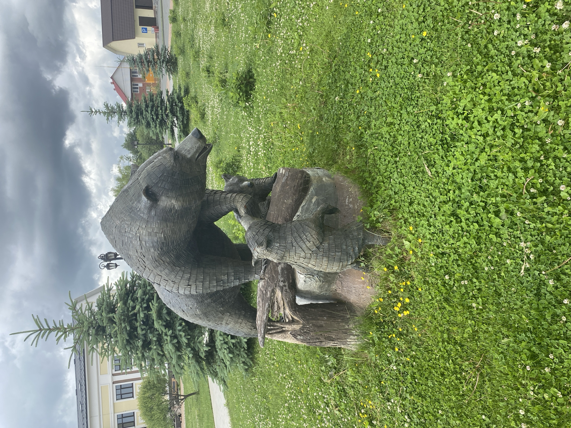 A dark metal sculpture depicting a mother bear with two cubs stands prominently on a rock and log base within a green park area in Tyumen, Russia. The mother bear, characterized by a textured, scaled surface, is positioned facing right, with one cub standing on its hind legs on the log, reaching towards her, and a second cub partially visible behind her. The surrounding ground is covered in dense green grass interspersed with white clover and small yellow wildflowers. In the left background, a light yellow building with dark windows and white trim is visible behind a tall evergreen tree, with a paved pathway in the immediate foreground. To the right of the sculpture, another evergreen tree precedes residential buildings with red and brown roofs; one yellow building on the far right displays a blue 'P' parking sign. A vintage-style lamppost with two lanterns is partially visible behind the statue. The entire scene is under a heavily overcast sky with dark gray clouds. No people are visible.