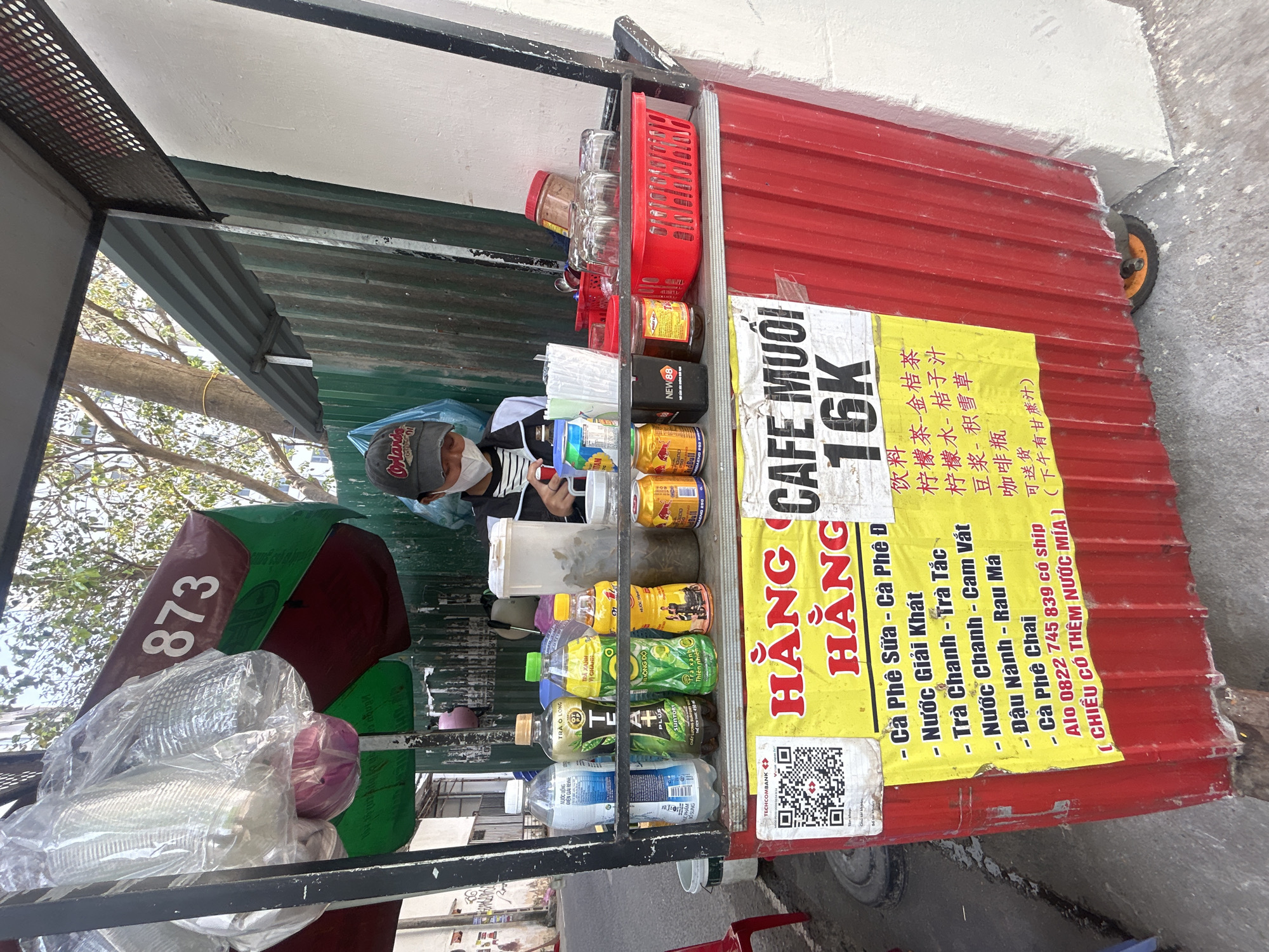 This image captures a vibrant street vendor stall in what appears to be Bắc Nha Trang, Vietnam. The stall, constructed from red corrugated metal, features a simple counter and a green corrugated roof for shade. A masked vendor, wearing a dark cap with white lettering, is partially visible behind the counter, seemingly attending to the stall or a device.
The stall is well-stocked with a variety of beverages. The upper shelf holds glasses in a red plastic basket and canned/bottled items, including brands like "NEW 88" and "RedBull." The main counter displays numerous bottled drinks such as "TRÀ Ô LONG," "SUNTORY TRÀ XANH," and "DRAGON GREEN TEA." Clear plastic cups and other supplies, wrapped in plastic, are stacked on the left. A large, folded green umbrella, partially displaying the number "873," leans against the left side of the stall.
Two prominent yellow signs are taped to the front of the counter. The larger sign advertises "CAFE MUỐI 16K" (Salt Coffee for 16,000 VND). Below this, a menu lists drinks in both Vietnamese and Chinese characters. The Vietnamese options include:
- Cà Phê Sữa - Cà Phê Đen (Milk Coffee - Black Coffee)
- Nước Giải Khát (Soft Drinks)
- Trà Chanh - Trà Tắc (Lemon Tea - Kumquat Tea)
- Nước Chanh - Cam Vắt (Lemon Juice - Orange Juice)
- Đậu Nành - Rau Má (Soy Milk - Pennywort Drink)
- Cà Phê Chai (Bottled Coffee)
The sign also notes, "Alo 0822 745 839 có ship" (Call 0822 745 839 for delivery) and "(CHIỀU CÓ THÊM NƯỚC MÍA)" (Sugarcane juice available in the afternoon). A QR code for "TECHCOMBANK" is visible on the left sign, indicating mobile payment availability.
The bright natural light and visible trees suggest a clear, daytime setting. A small wheel is visible on the right side of the stall, hinting at its mobility.