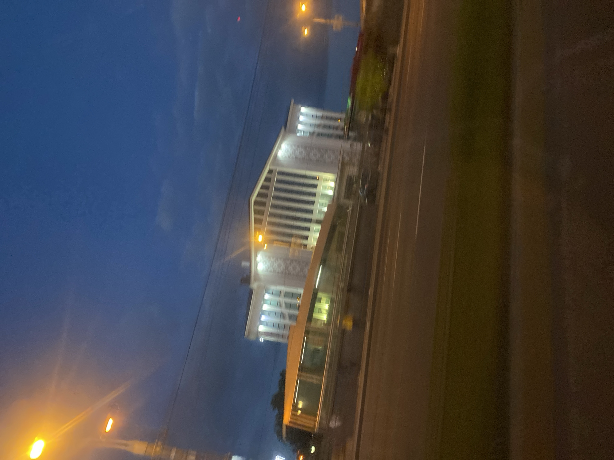 The image shows a contemporary building with a modern architectural style, likely in Minsk, Belarus, at dusk or early evening. The building features a prominent white facade with numerous vertical windows and decorative elements. A sloping glass-covered walkway or bridge connects to the building. In the foreground, a street with at least two lanes of traffic is visible, with a grassy area separating the road from the building. Streetlights with orange illumination are present, casting a warm glow on the scene. The sky is a deep blue with some clouds, indicating the transition from daylight to night. No people are clearly visible in the image, but a dark-colored car is parked near the building. The overall impression is one of urban infrastructure bathed in artificial light against a darkening sky.