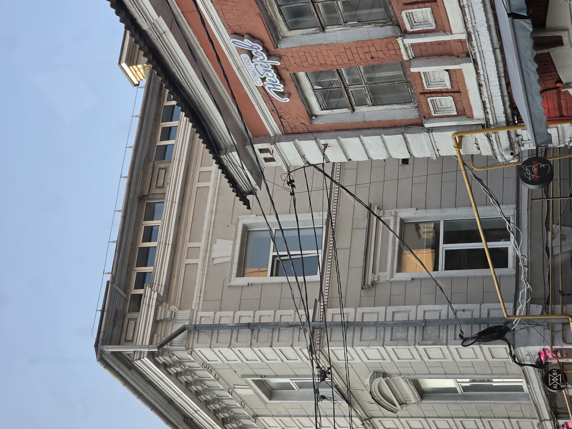 The image shows the facade of two adjacent buildings in Nizhny Novgorod, Russia, under a clear blue sky. The building on the left is a light-colored, multi-story structure with ornate architectural details, including a decorative cornice and pilasters. It features several large windows, some with white frames and others with reflections visible. A drainpipe runs vertically along the corner of this building.
The building on the right is constructed of red brick and has white decorative elements. It also has several windows, some of which appear to be old or in disrepair. A sign with white cursive lettering reading "СТОМАТОЛОГ Мещеряков" (Dentist Meshcheryakov) is mounted on the brick facade.
Numerous black electrical wires crisscross between the buildings, creating a complex network in the foreground. A black object, possibly a piece of electrical equipment or a decorative element, is attached to the drainpipe on the left building. A yellow pipe runs horizontally in the middle of the frame, and a gray awning is visible extending from the red brick building at the bottom. A round sign with a stylized face is also present near the yellow pipe. The overall impression is a view of urban architecture with visible infrastructure.