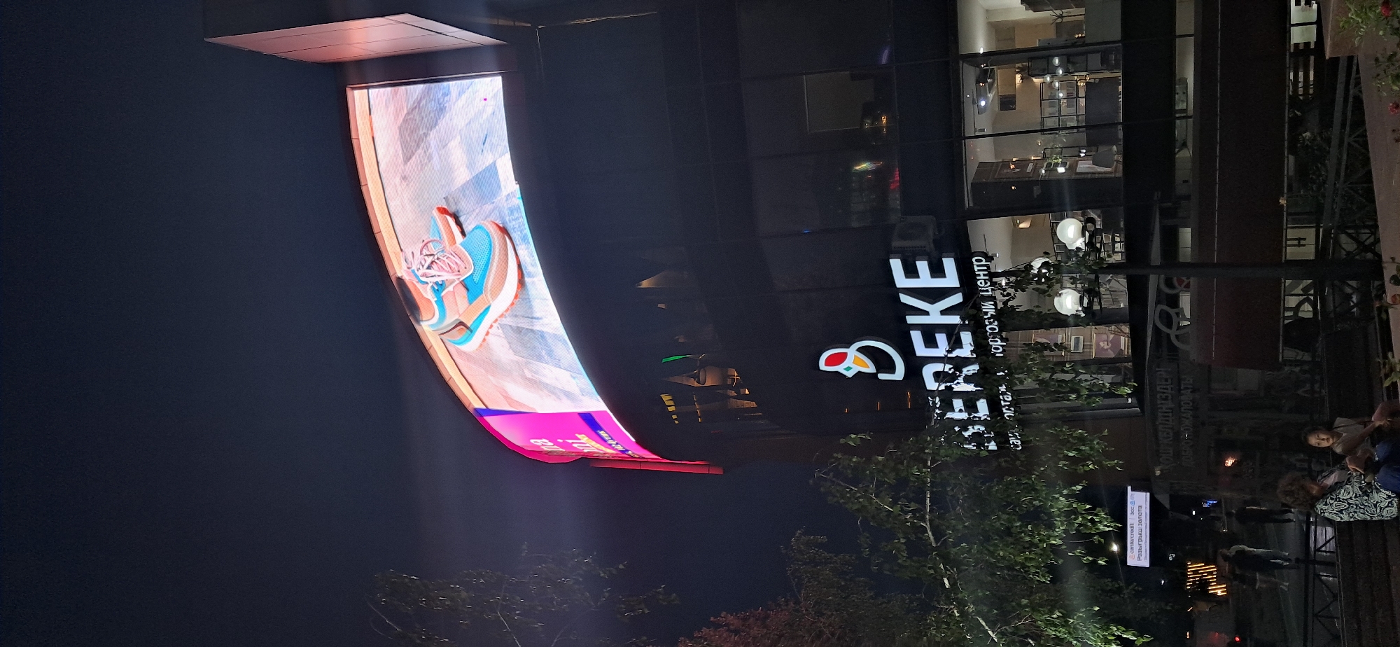 This media file depicts a nighttime scene outside a commercial building with a prominent digital billboard. The billboard, on the left side of the frame, displays an advertisement for what appears to be athletic shoes, featuring a close-up of a blue and pink sneaker. The billboard is curved and illuminated, casting a glow on the dark surroundings.
To the right of the billboard is the building itself, which has a dark facade with large glass windows. Through the windows, various internal lights and displays are visible, suggesting it is an active commercial space. A sign on the building reads "EREKE" in large white letters, accompanied by a logo that appears to be a stylized bird or flame. Below this, there is Russian text which is partially obscured and difficult to read, but appears to include "торговый центр" (shopping center).
In front of the building, there are trees with green foliage, and several streetlights with globe-shaped lamps. Some people are visible in the scene, including a group sitting on a bench on the right side of the frame, partially obscured by foliage. One person is looking towards the camera. The overall atmosphere is that of an urban evening, with artificial lights dominating the scene. The time of day is clearly night, and the weather appears to be clear.