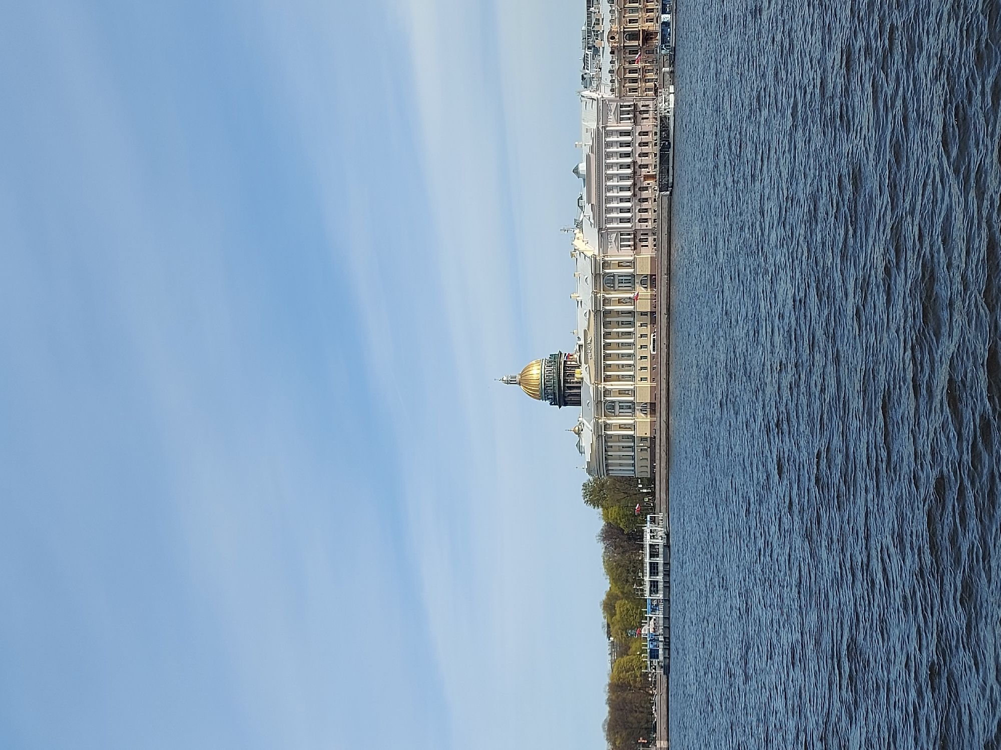The image depicts a waterside view of Saint Petersburg, Russia. On the right side is dark blue water, rippled by wind or current. To the left of the water is a portion of the city's riverbank. The architecture along the riverbank features several buildings, including one with a prominent golden dome. The dome suggests the presence of a cathedral or significant landmark. Trees line the edge of the riverbank. The sky in the background is light blue with streaks of white clouds. The scene is captured in daylight with good visibility. The photograph is taken from a lower-ground or boat-level perspective.