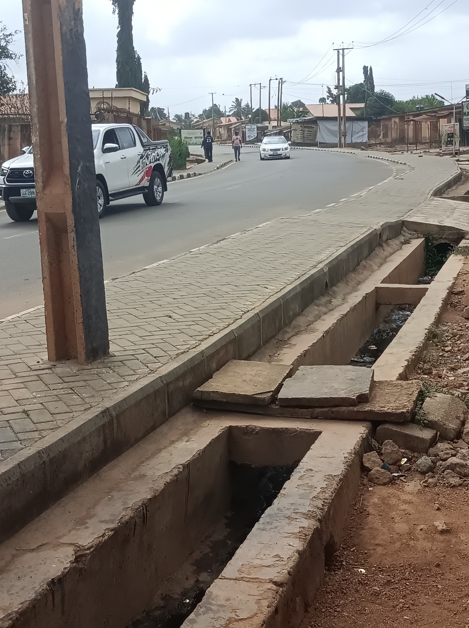 This media file captures a street scene in Narayi, Nigeria, on an overcast day. The image is dominated by a paved road curving to the right, bordered by a paved sidewalk constructed from grey rectangular bricks. Adjacent to the sidewalk is a concrete drainage ditch, partially covered with rough concrete slabs, revealing accumulated debris and stagnant water in the open sections.
In the foreground, a tall, rectangular pole, half brown and half black, stands prominently on the left. On the road, a white Toyota Hilux pickup truck with the license plate "GEE 20RN" and "INVINCIBLE" printed on its side is seen. Further down the curved road, a white sedan is visible, and a few individuals are walking, engaged in everyday activity.
The background features various low-rise buildings, some resembling market stalls or shops, with simple roofs. Utility poles with overhead lines crisscross the sky. To the right of the drainage ditch, the ground is rough with rocks and dirt. A partially visible sign in the distance advertises "SOYA OIL" and "YOGURT & P," suggesting local commerce. The overall scene depicts a typical urban or semi-urban street environment with a mix of infrastructure and daily life.