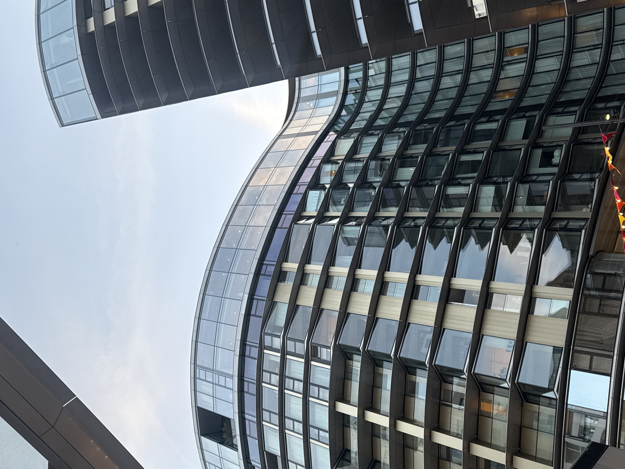 Here is a description of the image:
The image shows a view of two modern buildings in what appears to be the city of London, United Kingdom. The right-most building has a distinctive curved shape and a facade composed of numerous glass windows with dark metal frames. Some windows are slightly ajar. The exterior surface is interrupted by horizontal bands, maybe of a lighter metal. The left-most building is a blocky dark glass tower. The perspective is from ground level, looking upwards towards the buildings and the sky. The sky is pale blue with some thin clouds. A red banner with flags is visible attached to the right-most building.
