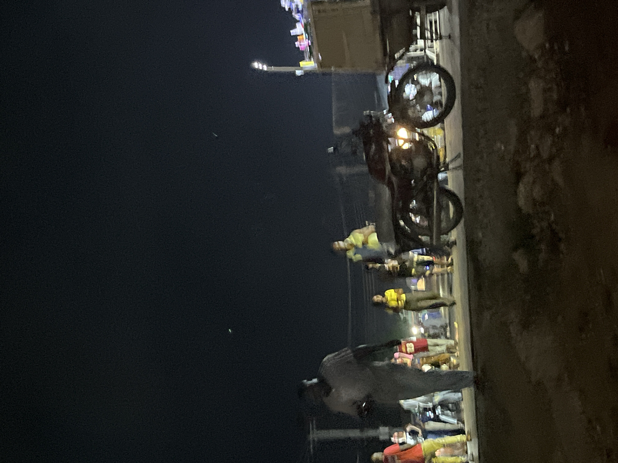 An outdoor, nighttime scene depicts a group of people on a paved surface, possibly a road or sidewalk, in an urban environment. Several individuals are visible, some walking and others standing. A motorcycle is parked in the foreground, with its headlight illuminated. In the background, a vehicle with a trailer is visible, adorned with colorful items. The sky is dark, indicating nighttime, with two faint green streaks, possibly indicative of light trails or atmospheric phenomena. The overall impression is one of a busy street scene at night.
