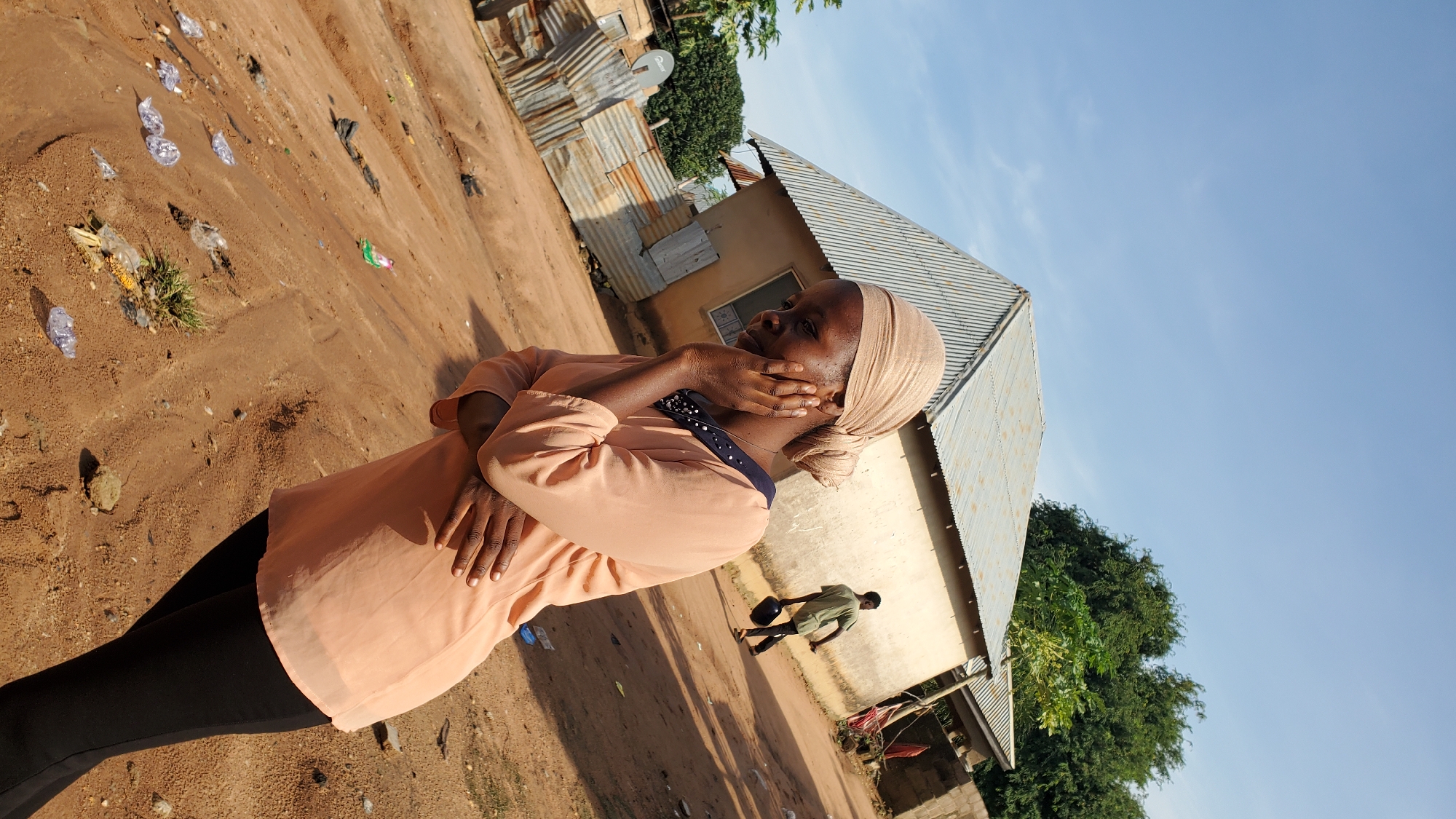 The image depicts an outdoor scene in Jalingo, Nigeria. In the foreground, a young woman wearing a peach-colored blouse and black pants is positioned on the right side of the frame, looking down and to her left with her right hand touching her face. Her arms are crossed in front of her. Behind her, a dirt path is visible, littered with plastic bottles and other debris. In the background, two buildings with corrugated metal roofs are present. One building appears to be a house, and next to it, a smaller structure with corrugated metal siding. Further in the background, a man in a green shirt and black pants is walking away from the camera, carrying a dark object, possibly a helmet, in his right hand. The sky is clear blue with some scattered clouds. Trees are visible in the upper right and left portions of the image. The overall environment suggests a semi-urban or rural setting.