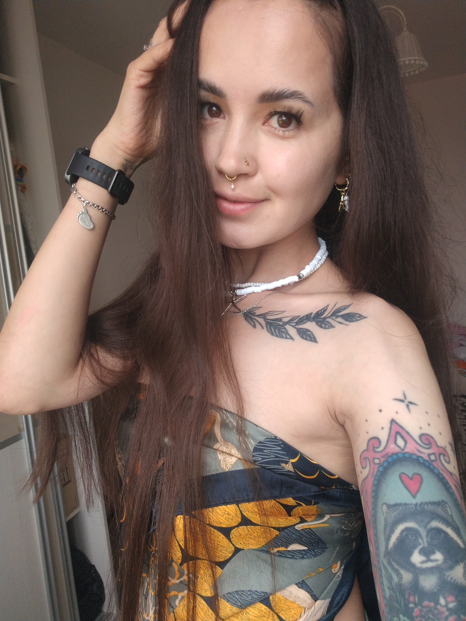 In the city of Noviy, Russia, a young woman is captured in a medium shot, posing for a selfie indoors. She has long, dark brown hair, a nose ring with a small diamond, and a gold hoop earring with a small charm. She is wearing a white beaded necklace and a black smartwatch on her left wrist with a silver bracelet featuring a small footprint charm. A tattoo of a branch with leaves is visible on her chest. She is also wearing a patterned top with blue and yellow floral designs. Her right arm is adorned with a colorful tattoo of a raccoon inside a pink-bordered frame, with a small red heart above it, and what appears to be a crown or star motif above that. The background is slightly blurred, showing what looks like a closet door and a lamp. The lighting suggests it is daytime.