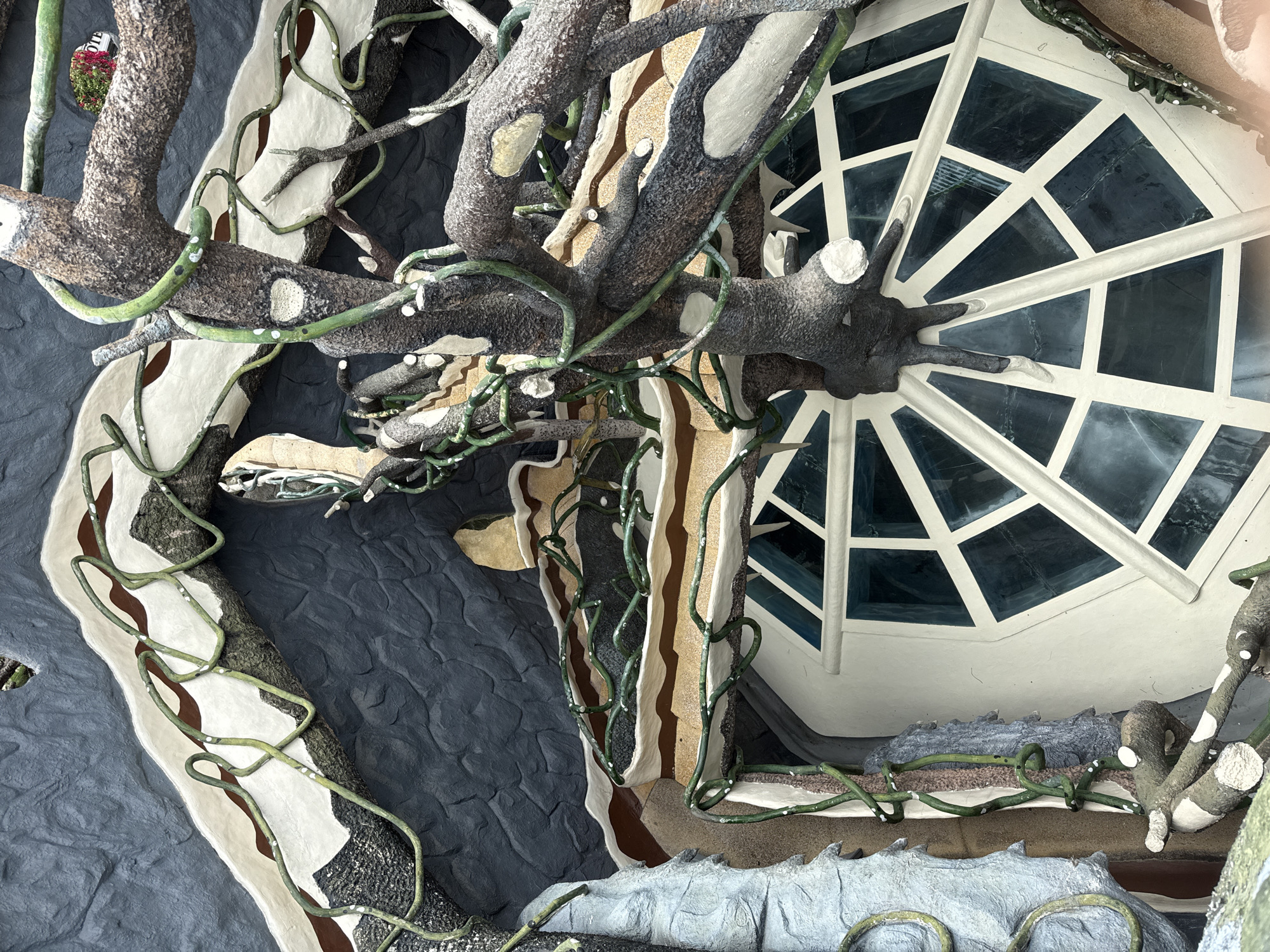 This image showcases architectural and decorative elements, seemingly part of a building's exterior, possibly in Da Lat, Vietnam. The design features faux-natural elements, including sculpted tree branches with white markings and intertwined green vines. A hexagonal window with geometric white framework is visible. The surrounding surfaces are sculpted to resemble layered earth or rock formations in shades of gray, beige, and brown, some with a textured surface. The style is unconventional and whimsical, reminiscent of art installations or highly stylized architecture.