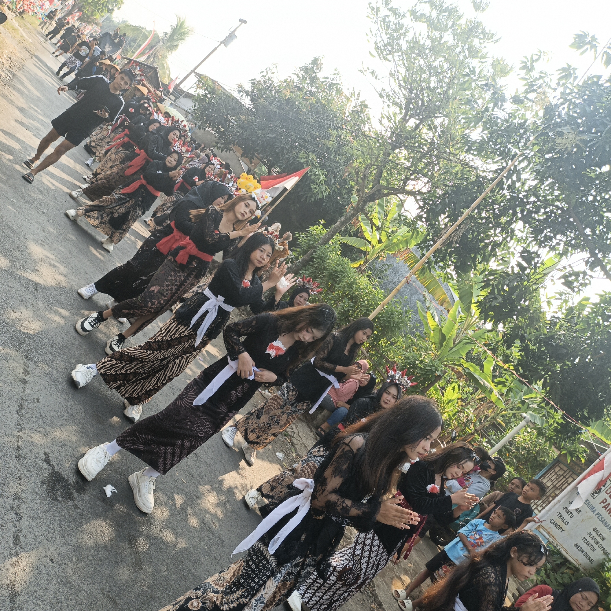 The image depicts a procession or performance on a paved street in Tanahbaya, Indonesia. A group of young women, dressed in traditional batik skirts and black lace tops with red or white sashes, are the primary subjects. Some wear ornate headpieces. They are in motion, with many having their hands clasped together in front of them. They are accompanied by onlookers, including children and adults, some of whom are holding Indonesian flags. The environment includes trees and buildings lining the street, and the scene is illuminated by natural daylight.