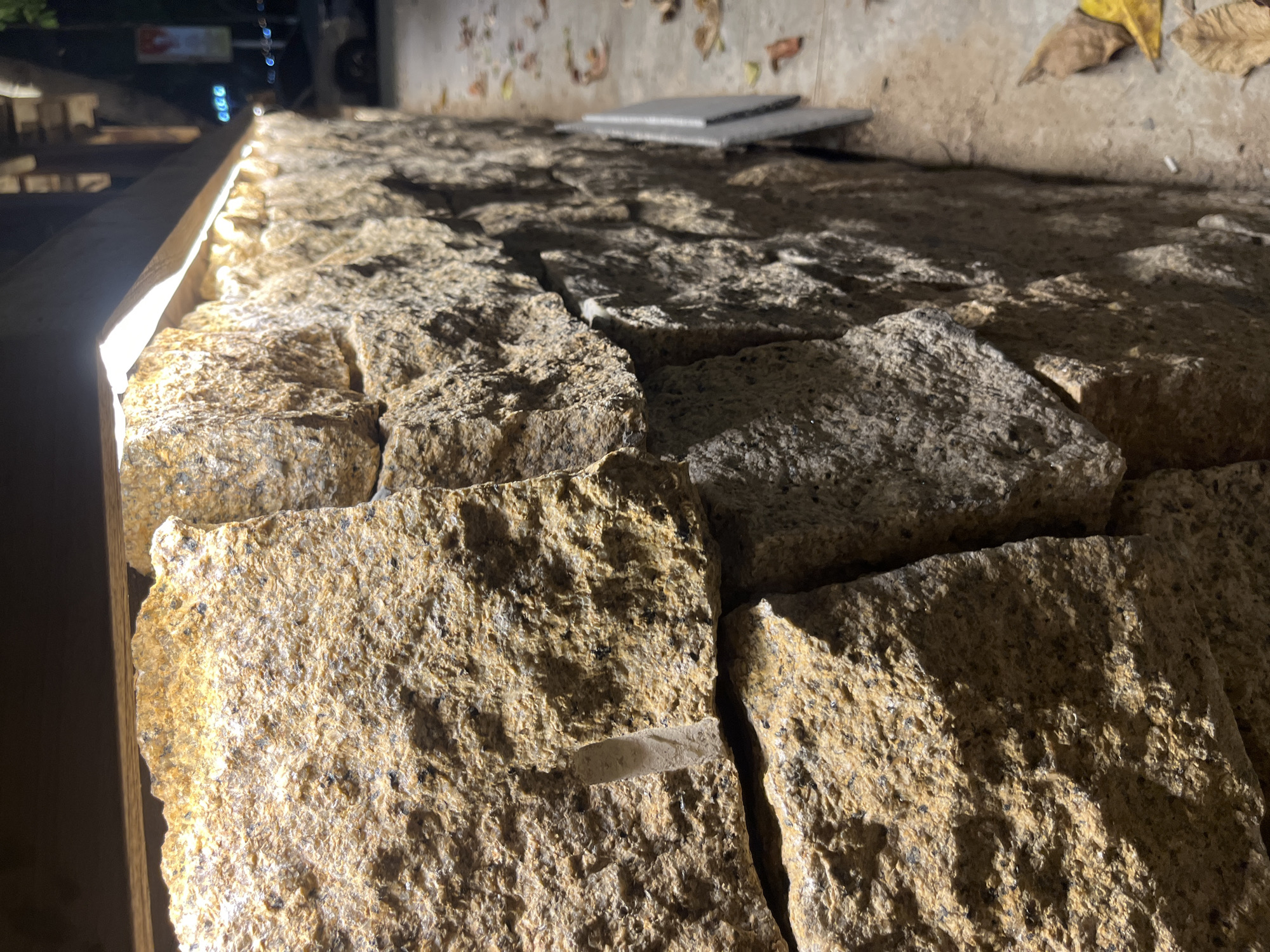 The image shows a close-up of a textured stone wall in Bac Nha Trang Ward, Vietnam, illuminated by a strip of LED lights positioned above the wall. The stones are irregular in shape and size, with a rough, granular surface exhibiting variations in color, primarily shades of tan and brown, speckled with darker mineral deposits. The lighting creates strong highlights and shadows, emphasizing the three-dimensional texture of the rocks.
To the right of the wall, a paved walkway is visible with scattered dry leaves. In the background, the scene is blurred, suggesting a nighttime setting. Streetlights and the lights of a building or vehicle are faintly discernible, along with the outline of a car. The overall impression is an outdoor urban environment at night, where the stone wall serves as a prominent architectural element. There are no people or discernible actions taking place in the image.