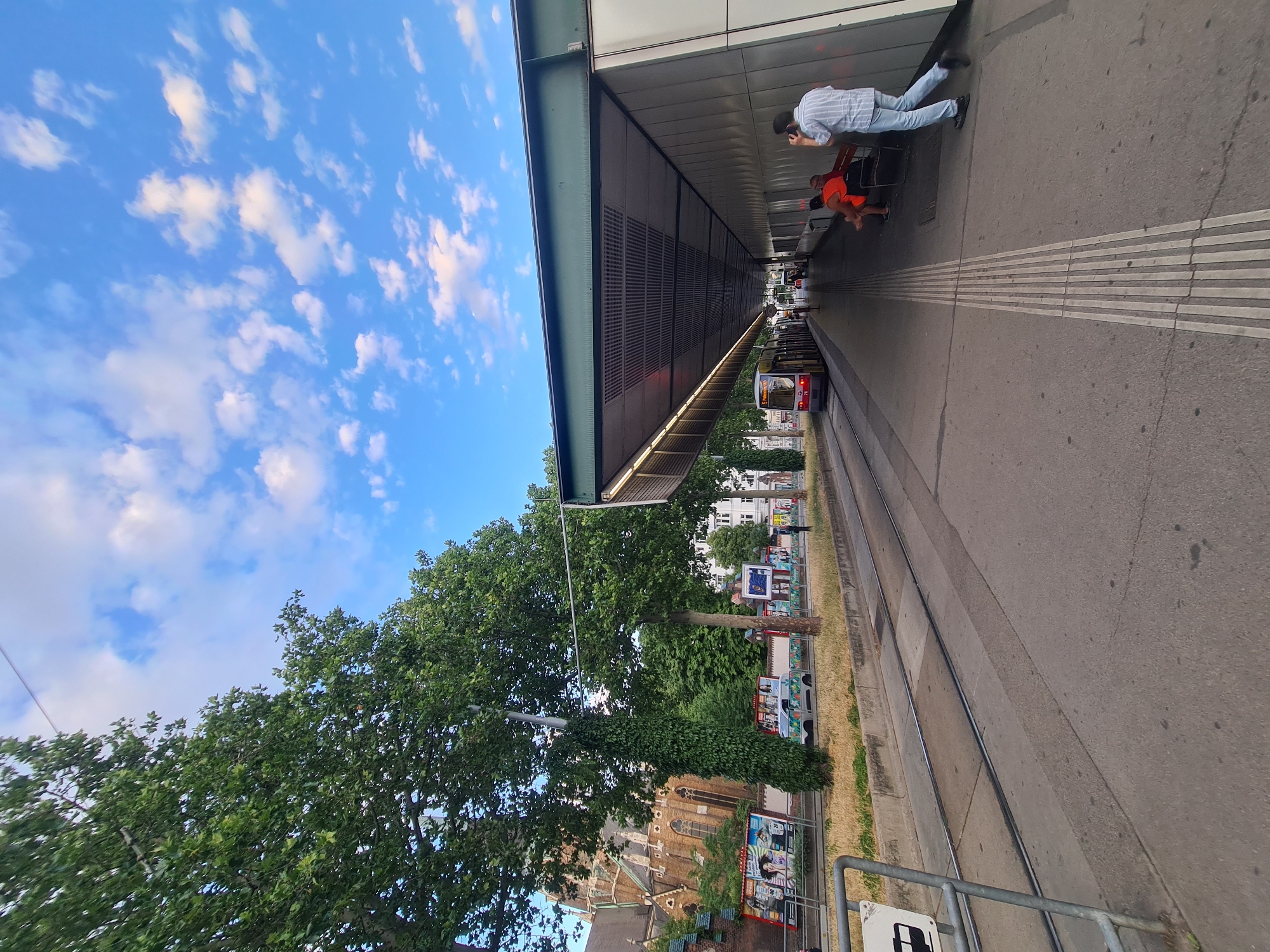 An outdoor, eye-level, wide shot shows a tram stop in Vienna, Austria. The tram stop has a modern canopy structure with a dark green roof and light grey walls. A tram, with the number 76 visible on its front, is stopped at the platform, which is marked with tactile paving for the visually impaired. Several people are seated on a bench under the canopy, and one person is standing, appearing to take a call. To the left of the tram tracks, a church with a prominent spire and ornate architecture is visible, suggesting an urban environment. Lush green trees line the street, and a bright blue sky with scattered white clouds forms the background. Advertisements are displayed on hoardings near the church. The overall scene depicts a functional public transport stop within a European city.
