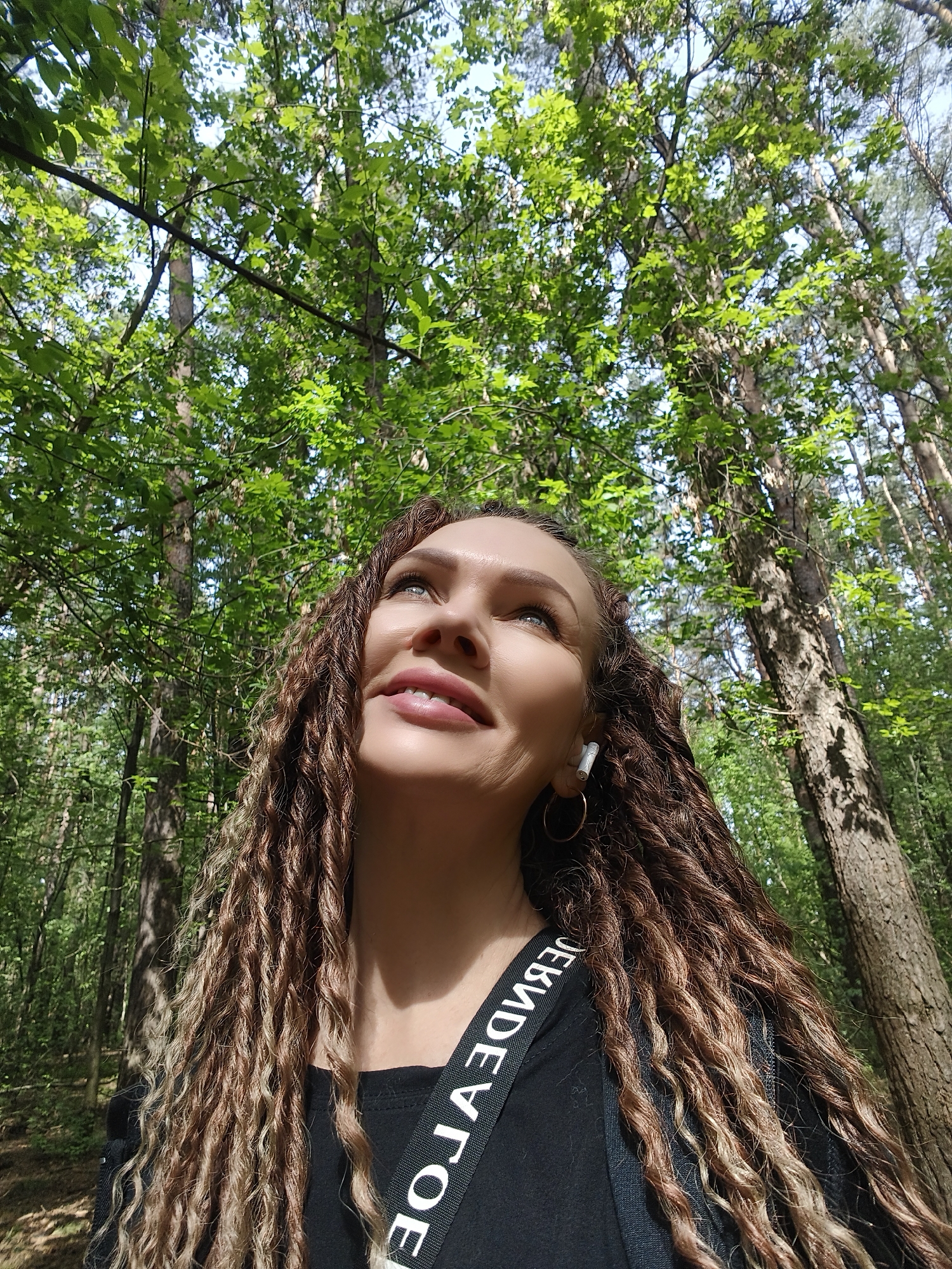 A low-angle medium shot captures a woman with long, braided hair, an earbud in her left ear, and a black top with a white strap over her shoulder, looking up towards the sky. She is positioned in a forest, surrounded by tall trees with green leaves. The location is Unknown, Unknown.