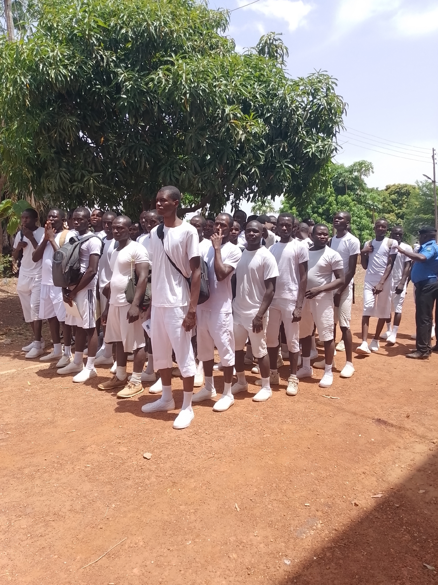 A group of young men, predominantly of African descent, are gathered outdoors under bright daylight. Most of the men are dressed in matching white t-shirts, white shorts, white socks, and white athletic shoes or sneakers, suggesting a uniform or dress code for an event. Several individuals are wearing backpacks, some dark, some lighter in color, and a few are holding papers or notebooks.
They are standing in what appears to be a line or formation on reddish-brown dirt or gravel ground. A large, lush green tree with dense foliage, possibly a mango tree, forms a prominent background, with a bright, partly cloudy sky visible above it. In the background to the right, utility poles and power lines are visible, indicating some infrastructure.
On the far right of the frame, a man in a blue short-sleeved shirt, dark trousers, and a dark cap (likely an instructor or official) is facing the group and appears to be speaking or giving instructions, gesturing with his right hand. The young men generally maintain attentive or neutral expressions, looking forward or towards the man in blue. The scene suggests an outdoor recruitment, training, or orientation session for a group like a police force, military, or a youth corps, held in a warm climate. Shadows indicate it is likely midday or early afternoon. No visible text is discernible.