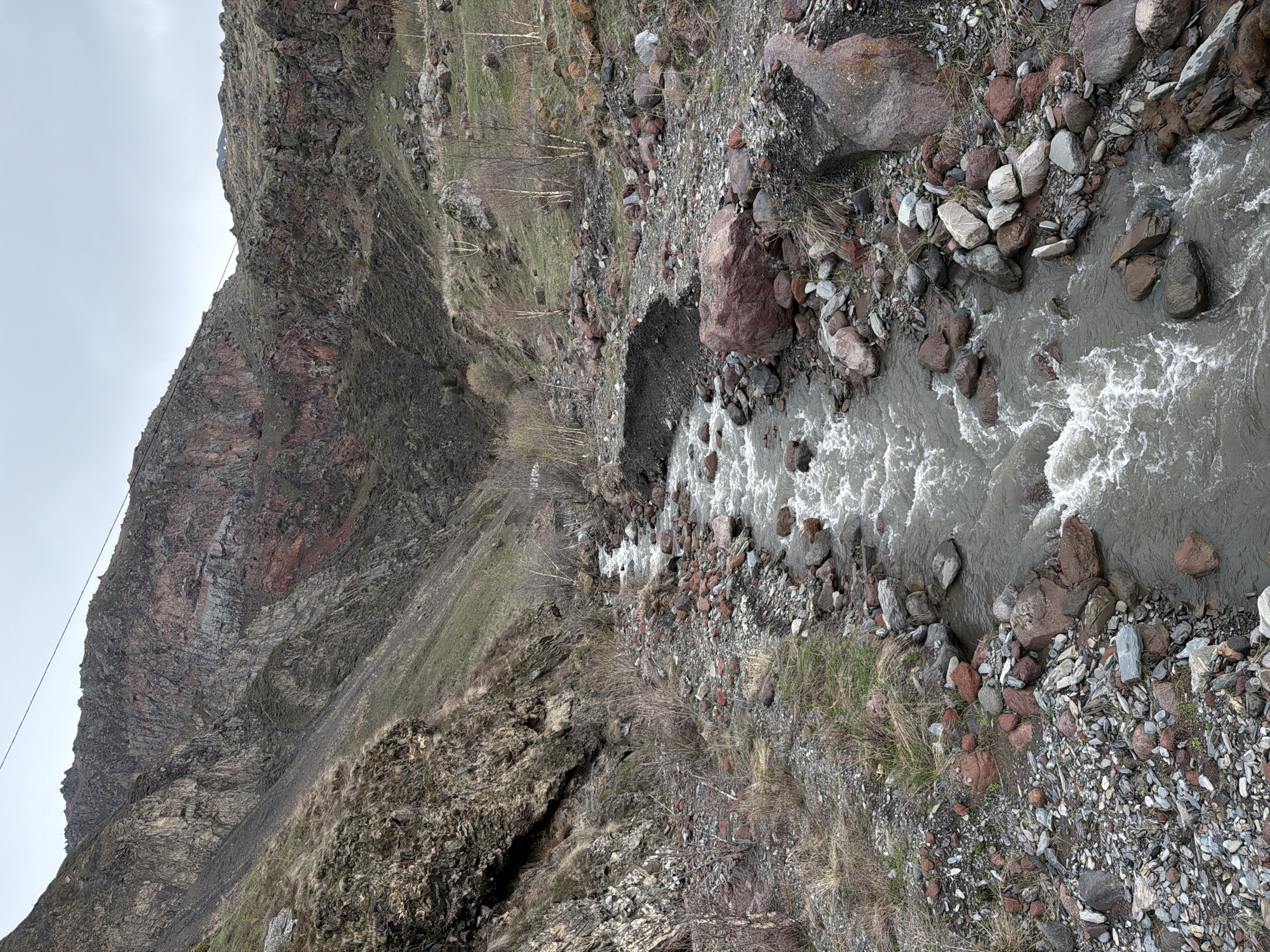 The image depicts a mountainous landscape with a river flowing through a rocky terrain. The location appears to be near Khurtisi, Georgia, based on the landscape features. A steep, rugged mountain occupies the left side of the image, exhibiting stratified rock formations and sparse vegetation. The riverbed is filled with various sized rocks and stones, with the river water appearing murky. There are no visible people or animals.