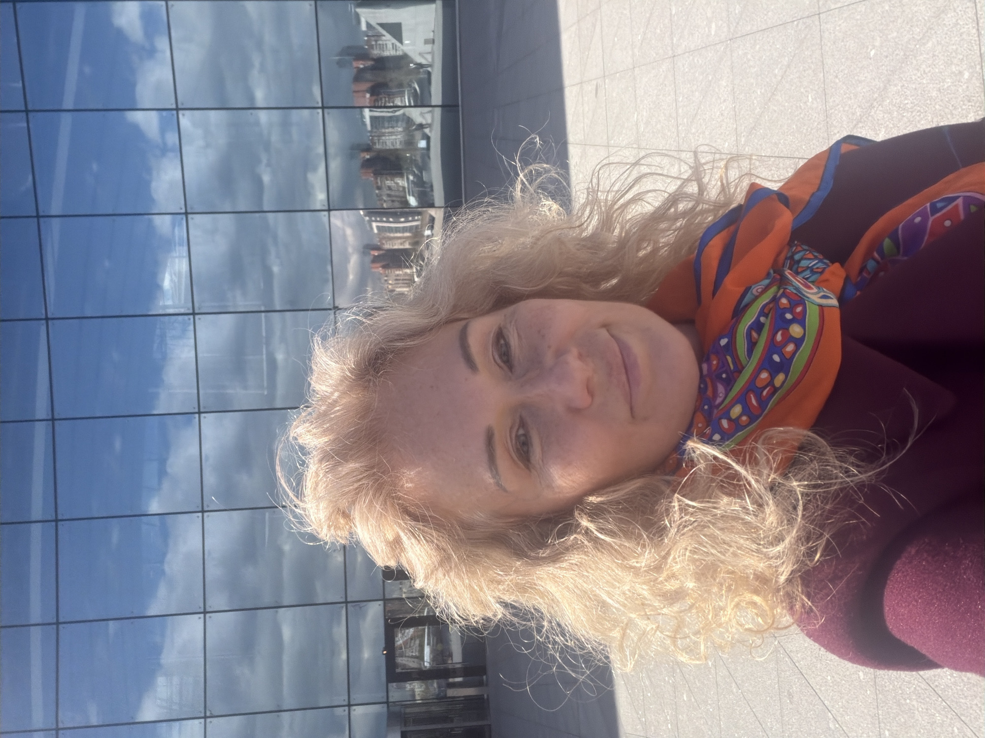 A woman with blonde curly hair, wearing an orange and blue scarf and a maroon coat, is taking a selfie outdoors. The background features a modern building with a glass facade reflecting the sky and other buildings. The ground is paved. Considering the architectural style reflected in the glass facade, the location is likely in a modern area of Saint Petersburg, Russia, but without additional context, specific identification is not possible.