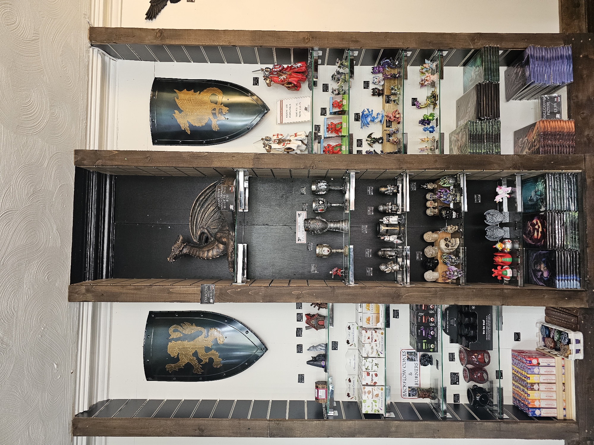 A retail display within a shop in Llandudno Junction, United Kingdom, features three rows of wooden shelves mounted on a wall. The shelves contain an assortment of fantasy and occult-themed items. The top shelf includes toy knights on horseback, small dragon figurines, and small canvas pictures. The middle shelf displays dragon statuettes, goblets, skulls, and fantasy-themed pictures. The bottom shelf contains shields with dragon emblems, incense cones, backflow burners, and manga books. Signs with prices are visible throughout the display.