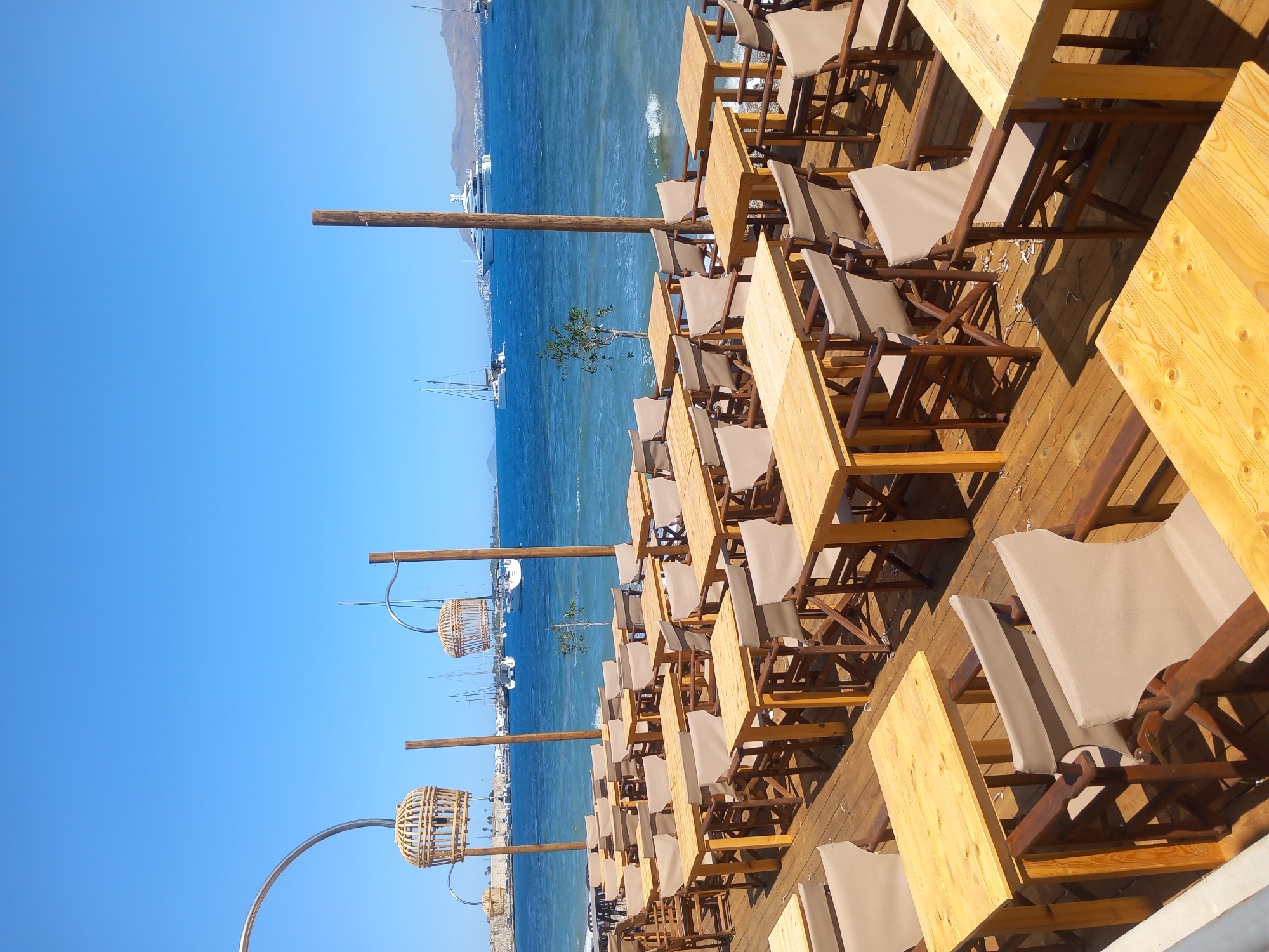 The image shows an outdoor seating area of a restaurant or cafe on a sunny day. The seating area is on a wooden deck overlooking a blue sea. Several tables and chairs are arranged on the deck. The tables are made of light-colored wood, and the chairs are made of wood with light-colored fabric seats and backs. In the background, there are sailboats and yachts on the water, and land with buildings can be seen in the distance. Two decorative bamboo bird cages hang from wooden poles. The location is identified as the city of Kos, Greece, and the scene appears to be a waterfront establishment. There are no people visible in the image. The weather is clear with a bright blue sky.