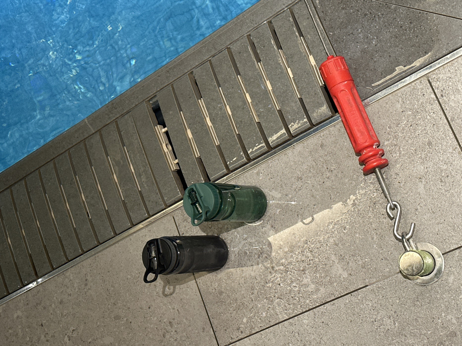 A pool's edge shows blue water next to a gray tiled border. A drainage grate runs along the border. Two water bottles, one dark and one green, lay on the tiles. A red-handled tool with a hook and metal attachment also sits on the tiles, near a drain cover. The scene appears to be at a pool.