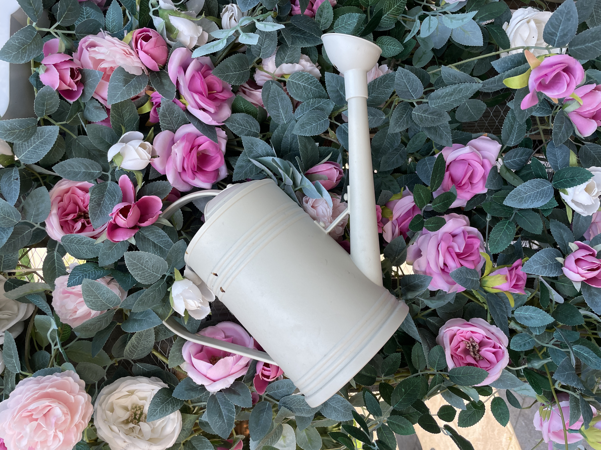 A cream-colored watering can is positioned diagonally across a dense arrangement of artificial pink and white roses and green foliage. The watering can's handle is visible on the left, and its spout extends to the right. The flowers and leaves form a lush backdrop, with many roses in various stages of bloom and tightly packed green leaves. The scene appears to be an outdoor decoration, possibly in a garden or public space.