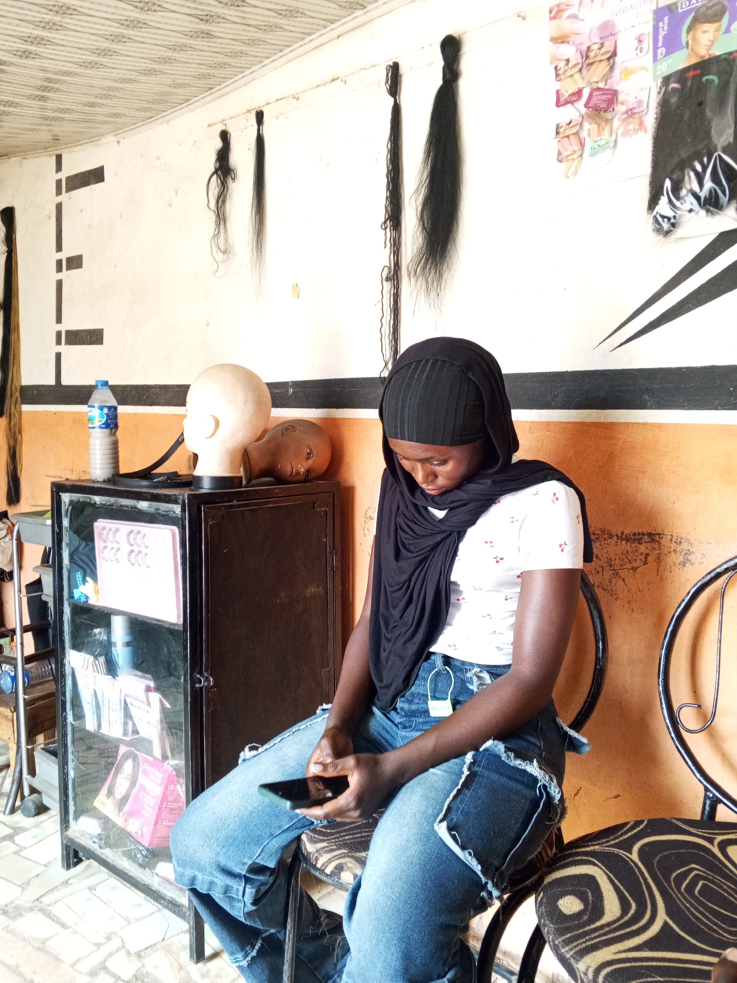 A young woman in a black headscarf and a white t-shirt with red cherries print is sitting on a chair in a small room in Gwong, Nigeria. She is wearing blue jeans with frayed edges and a dark scarf draped around her neck. She is looking down at a smartphone she holds in her hands.
The room appears to be a small hair salon. Behind her are walls with orange and white paint, adorned with black lines. Mannequin heads are on top of a black metal cabinet, with hair extensions hanging on the wall. The cabinet contains various hair and beauty products. A water bottle sits on top of the cabinet. A second chair with a patterned seat is next to the one she occupies. The floor is tiled in a light gray pattern.