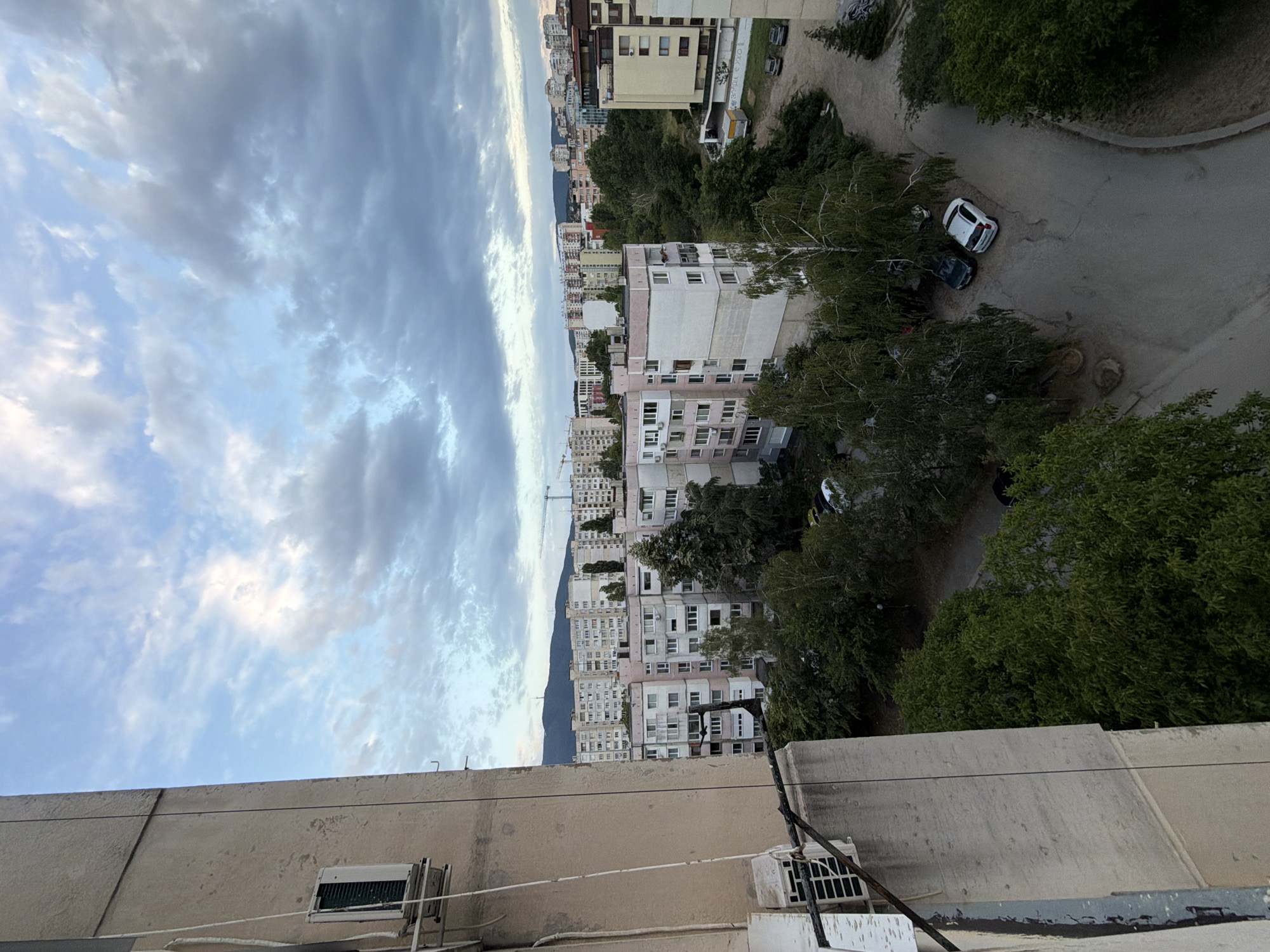 The image captures an aerial view of a residential area in Sofia, Bulgaria, likely taken from a balcony of a high-rise building. The foreground features the weathered concrete facade of a building with an external air conditioning unit attached. To the right, the scene opens up to a panorama of Soviet-era apartment blocks, characterized by their pastel-colored facades and repetitive window patterns. Some buildings are pink and white, while others are a pale yellow. Lush green trees and foliage are interspersed between the buildings, suggesting a relatively well-maintained urban green space. A network of asphalt roads and parking areas is visible, with a few cars parked in open spaces. In the distance, a hazy silhouette of mountains can be seen, typical of the Sofia basin's geography. The sky is partly cloudy with dramatic, overcast formations, indicating either early morning or late afternoon light. Construction cranes are also visible in the background, hinting at ongoing development within the city. There are no people or discernible human activity in the frame. The overall impression is one of a dense, established urban neighborhood with a blend of architecture and natural elements.