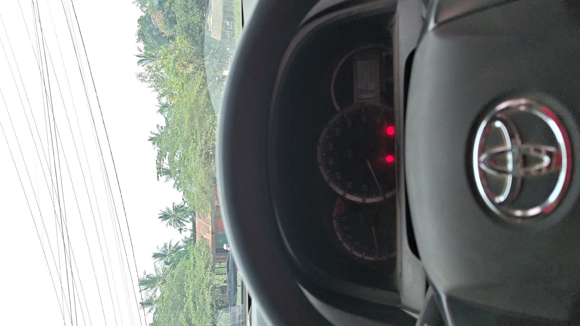 The image is a first-person perspective from inside a Toyota vehicle, looking forward through the windshield. The steering wheel, with the Toyota logo prominently displayed in the center, is in the foreground. The dashboard features a speedometer and other gauges, with two red indicator lights illuminated. Outside the vehicle, a bright, overcast sky is visible, with a series of power lines crisscrossing the upper portion of the frame. Below the power lines, lush green foliage, including palm trees and other vegetation, suggests a tropical environment. Several simple buildings, some with corrugated metal roofs, are nestled amongst the trees, indicative of a rural or suburban setting in Manetunong, Indonesia. The scene appears to be captured during daylight hours.