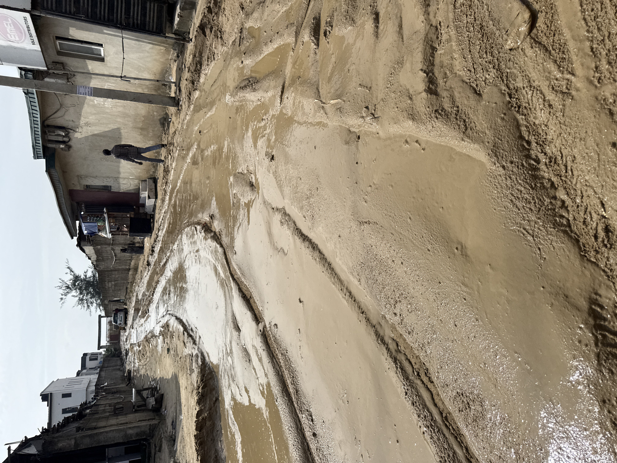 The image depicts a flooded street in Ado, Nigeria. The street is covered in thick, muddy water with visible tire tracks. A man is walking on the sidewalk next to a building with a sign that reads "EMEL SOLE DISTRIBUTORS". Other buildings and vehicles are visible in the background. The sky is overcast.