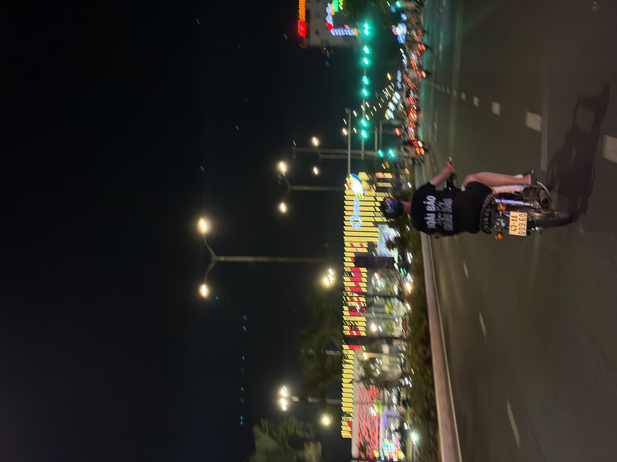 The image shows a night scene on a city street, likely in Đà Nẵng, Vietnam, with a person riding a motorcycle in the foreground, moving away from the camera. The rider is wearing a black t-shirt with text on the back and a helmet. The motorcycle has a yellow license plate with black lettering that reads "43-AA 009.60".
The street is illuminated by streetlights and the bright lights of buildings lining the road. In the background, there's a large building complex with decorative yellow and red lights, suggesting it might be a shopping center or entertainment venue. Other vehicles and motorcycles are visible on the road, indicating traffic. The sky is dark, typical of nighttime. The overall impression is a busy urban environment at night, possibly in a commercial or entertainment district.