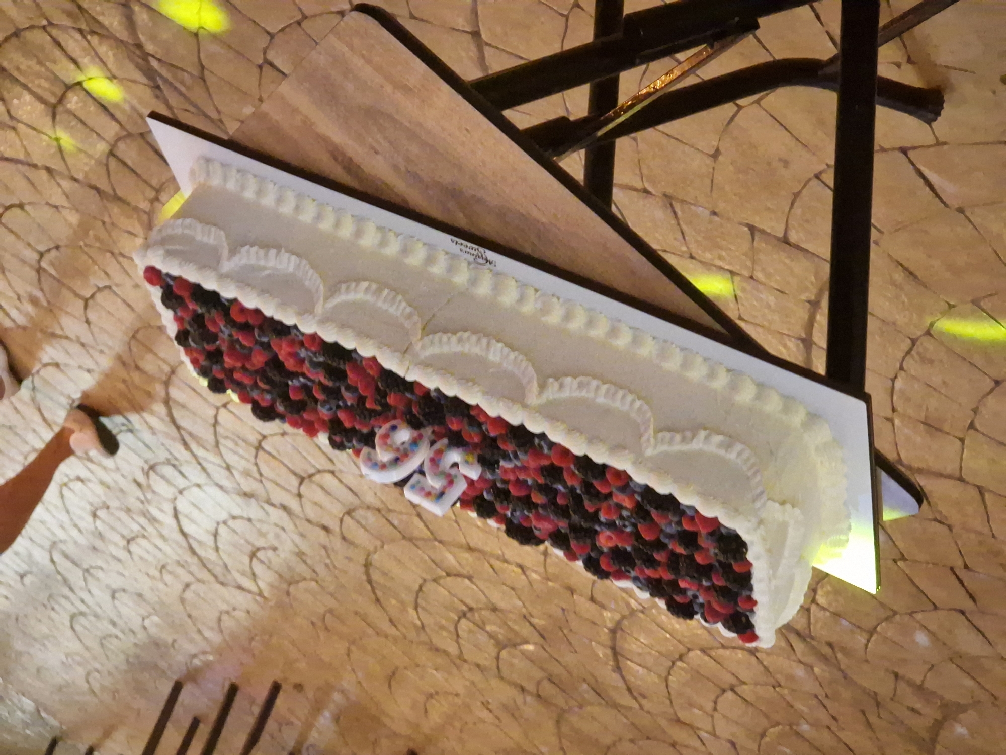 A long, rectangular cake adorned with a generous topping of fresh raspberries and blueberries sits on a tiered stand. The cake is covered in white frosting, with decorative piping along the edges. Two candles, shaped like the numbers "5" and "6", are placed on the berries, indicating a 56th birthday celebration. The cake is positioned on a wooden table with metal legs. The background features a patterned floor with mosaic-like tiles and some scattered yellow lights, suggesting an indoor or covered outdoor event space. A person's leg and foot, wearing a white sneaker, are visible on the right side of the image, hinting at the presence of guests. The scene is likely set in Tankovo, Bulgaria, given the context provided.