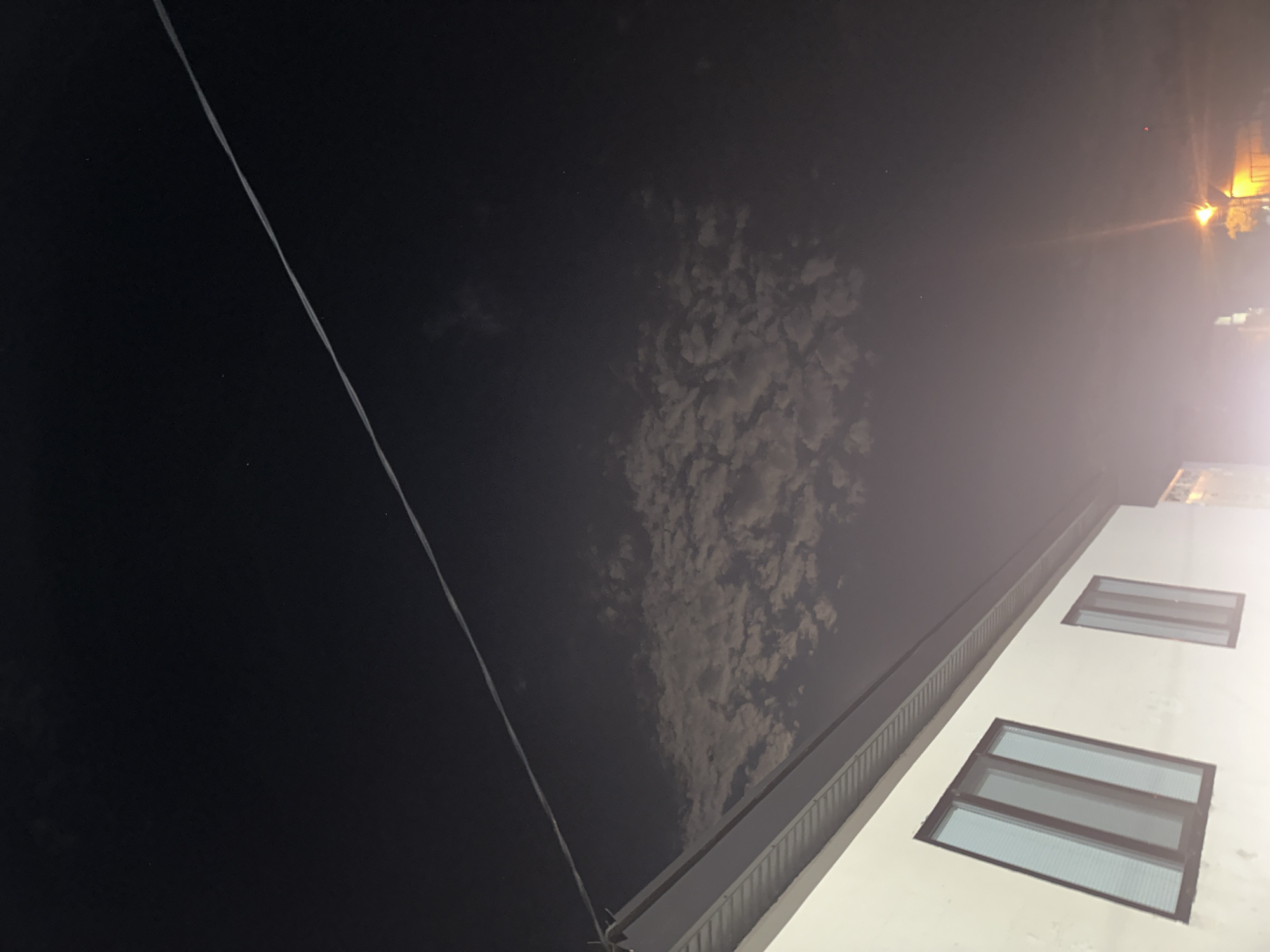 The image captures a nighttime scene in Subutli-Miatli, Russia. A portion of a white building with two windows is visible in the lower left, with a dark roofline extending diagonally across the frame. Above the building, a thin, white electrical wire stretches across the dark sky. Scattered, wispy clouds are illuminated faintly in the mid-sky, suggesting some atmospheric diffusion of light. In the lower right, a bright, orange street light casts a glow, and some indistinct structures are visible behind it. The overall atmosphere is dark and slightly hazy.