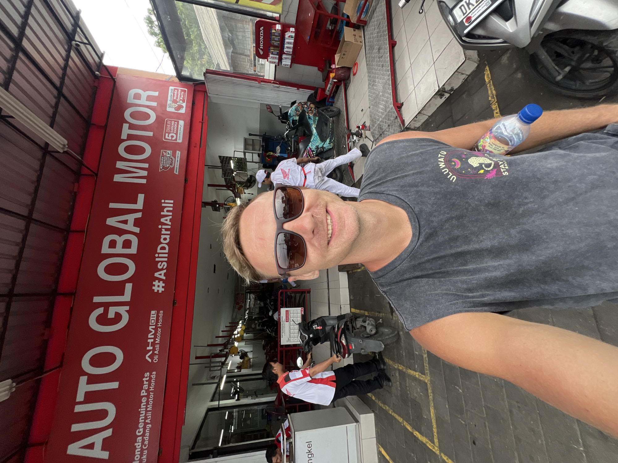 A man is taking a selfie in front of an Auto Global Motor Honda repair shop in Kuta Selatan, Indonesia. The man is wearing sunglasses and a grey t-shirt with an “Uluwatu” logo on the sleeve, and holding a water bottle. The shop has a large red sign with white lettering. Inside, mechanics wearing white and red uniforms work on scooters. A silver scooter is parked near the man.