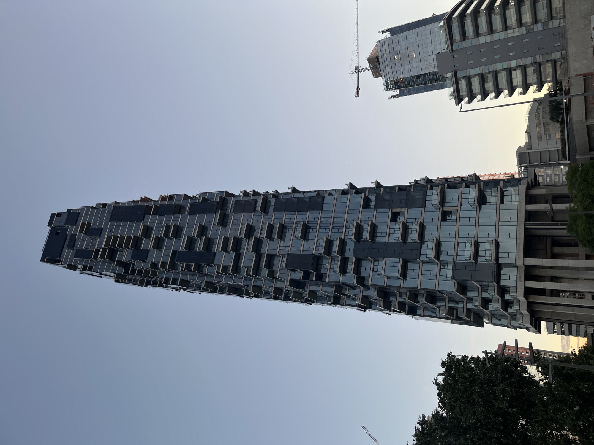 The image shows a tall, modern building with a distinctive facade of protruding rectangular balconies or extensions, located in Buenos Aires, Argentina. The building is primarily constructed of glass and dark gray materials. The sky is a pale blue. To the right of the primary building, another partially visible modern building under construction is seen, with a crane tower visible above it. In the foreground, the tops of trees are visible, suggesting the perspective is from a street level. The buildings' style and the urban environment suggest it's a modern residential or commercial district.