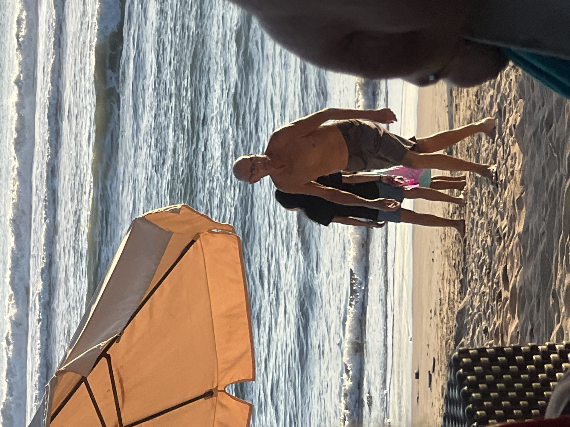 An older man with glasses and a bald head, wearing grey shorts and no shirt, walks on a sandy beach in Kuta, Indonesia. He is accompanied by two younger individuals, one wearing a black shirt and denim shorts, the other a blue shirt and shorts, carrying a pink bag. In the foreground, part of an orange umbrella and a black woven chair are visible. The ocean with waves is in the background, with a bright sky above.