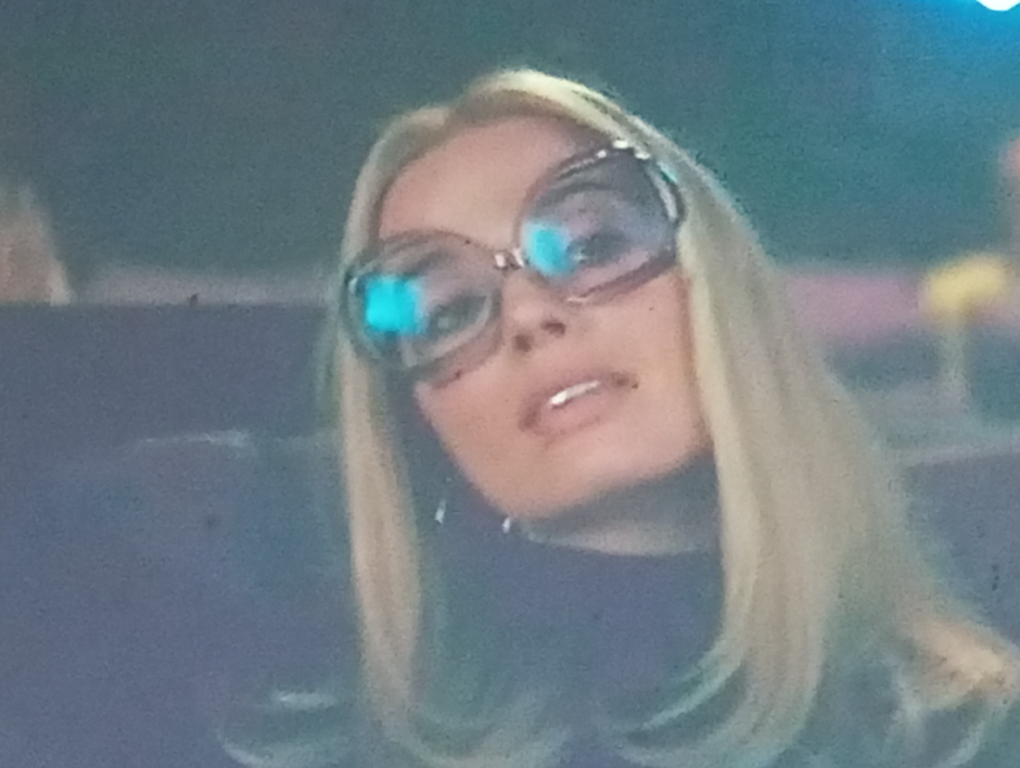 The media file is a close-up, medium-shot, slightly blurry photograph of a woman's face from the chest up. She has blonde hair styled straight and falling past her shoulders. The woman is wearing large, round, oversized glasses with thick, ornate frames that appear to be decorated with rhinestones or jewels. The lenses of the glasses are reflective, showing bright blue and white highlights, possibly from lighting in the environment. Her lips are slightly parted, and her expression is neutral or perhaps slightly pensive. She is wearing a dark-colored top, possibly a turtleneck. The background is out of focus, featuring muted blues and greens, with some indistinct shapes and colors that suggest an indoor or possibly an outdoor setting with soft lighting. There is no visible text. The overall impression is a portrait shot with a focus on the woman's face and her distinctive eyewear.