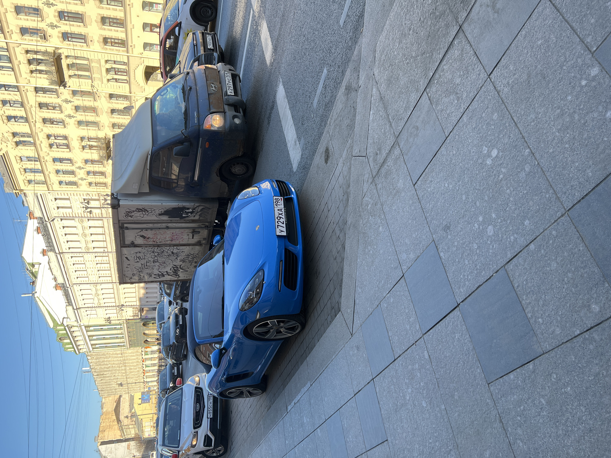 The image shows a street scene in Saint Petersburg, Russia. Cars are parked along the side of the road. A blue Porsche with license plate number У729ХА 198 is parked on the right side of the street. A blue van with license plate number А297РС 51 is parked in front of the Porsche. Another car with license plate number Н855СН 198 is parked behind the Porsche. In the background, buildings are visible, including one with ornate architectural details. The sky is blue and clear. The pavement is made of gray square tiles.