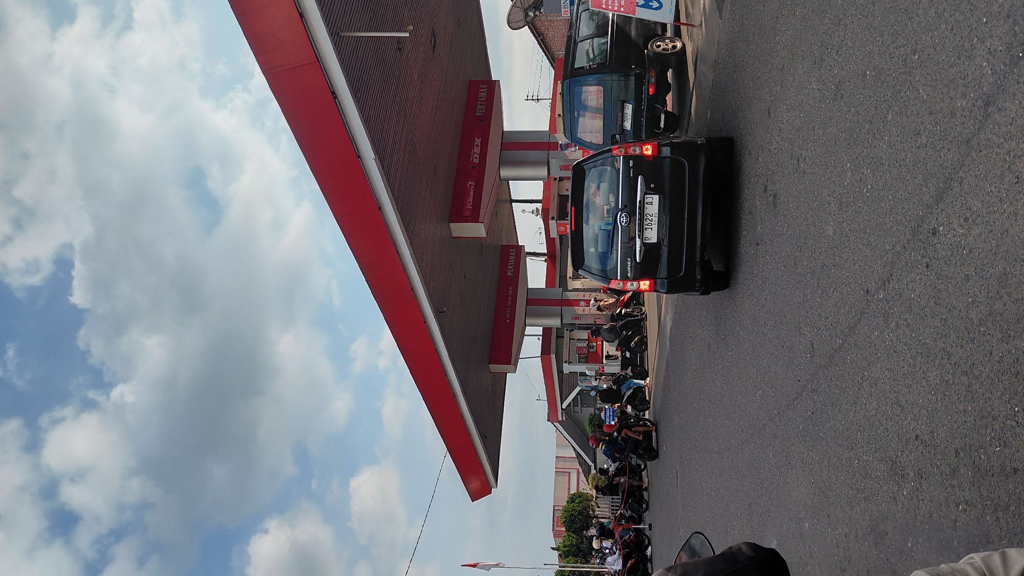 The image depicts a scene at a Pertamax gas station in Rejasari, Indonesia. In the foreground, a black Toyota Avanza with license plate R 1622 AJ is stopped on the asphalt road, facing away from the camera. To the right of the Avanza, a black utility vehicle is also parked. A red and white Pertamax gas station canopy extends over the vehicles and gas pumps. Several motorcycles with riders are lined up near the gas pumps, suggesting a queue for refueling. Some individuals are also standing near the motorcycles. In the background, buildings with red roofs are visible, along with a flagpole flying the Indonesian flag. The sky is partly cloudy with a mix of blue sky and white clouds. The overall scene suggests a typical day at a busy gas station in Indonesia.