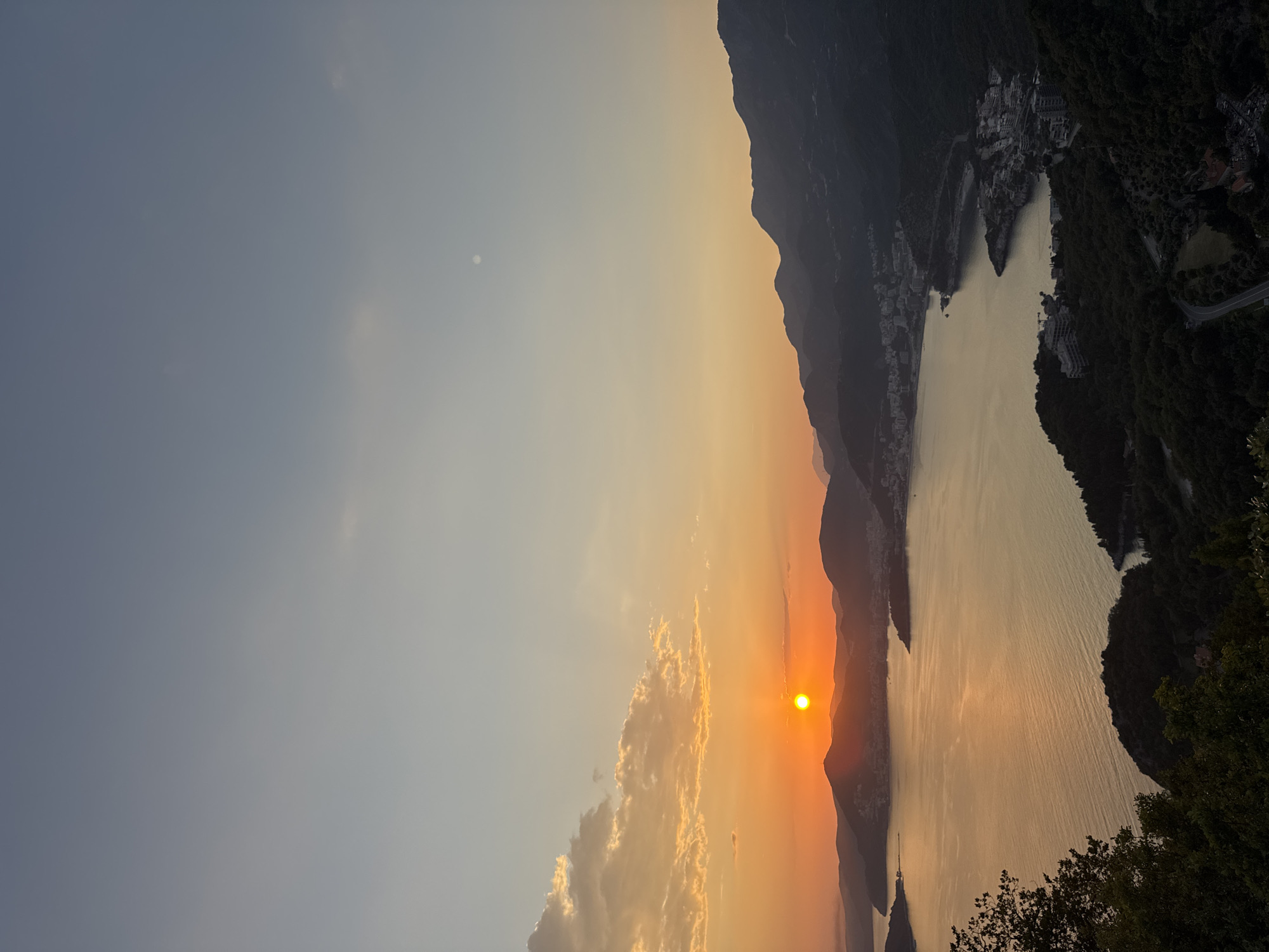 The image shows a sunset over a body of water, possibly the Adriatic Sea, in the vicinity of Sveti Stefan, Montenegro. The sun is setting behind mountains, casting an orange glow on the water and sky. The silhouette of mountains and coastal buildings is visible on the right side of the frame. The sky transitions from light blue to orange near the horizon. Some clouds are visible in the lower center of the image. A cluster of trees is visible at the bottom right.