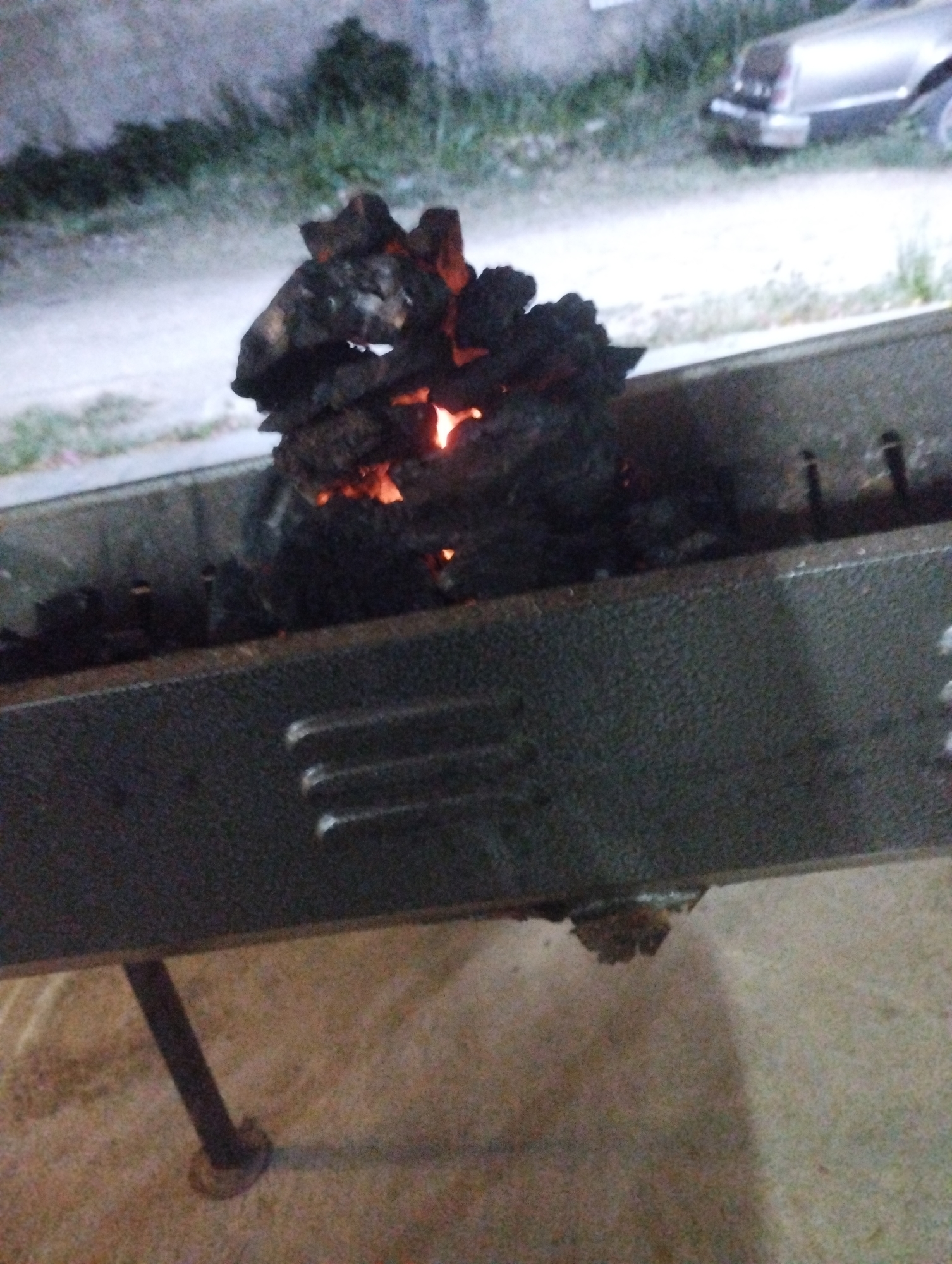 The image shows a charcoal grill with hot coals in Tinaquillo, Venezuela. The grill is grey and made of metal, with air vents on the front. It stands on a single leg, resting on a beige surface, possibly concrete or dirt. Hot coals are piled in the center of the grill, with visible embers glowing. In the background, there is a paved area with some vegetation and a portion of a car. The image appears to be taken at dusk or night.