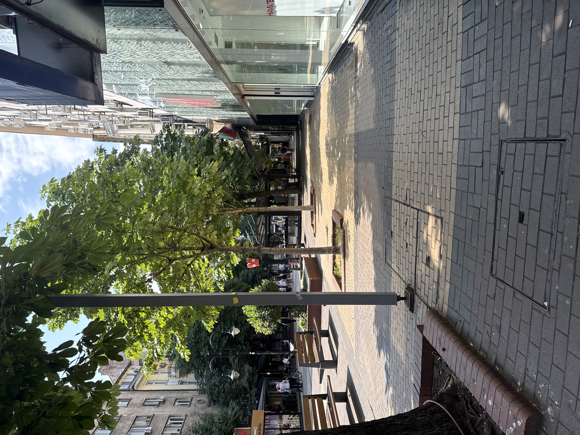 The image shows a street scene with a sidewalk lined with trees and benches. People are walking along the sidewalk and sitting on benches. Buildings with shops and windows are visible on one side of the street. The sun is shining, casting shadows on the sidewalk. The time of day appears to be daytime, likely late morning or early afternoon, given the angle of the sun. The weather seems pleasant. There are several trees providing shade, and the sidewalk is paved with bricks. Some of the shops have awnings or covered outdoor seating areas. One shop has a sign that reads "hamam". The overall atmosphere appears to be relaxed and urban.
