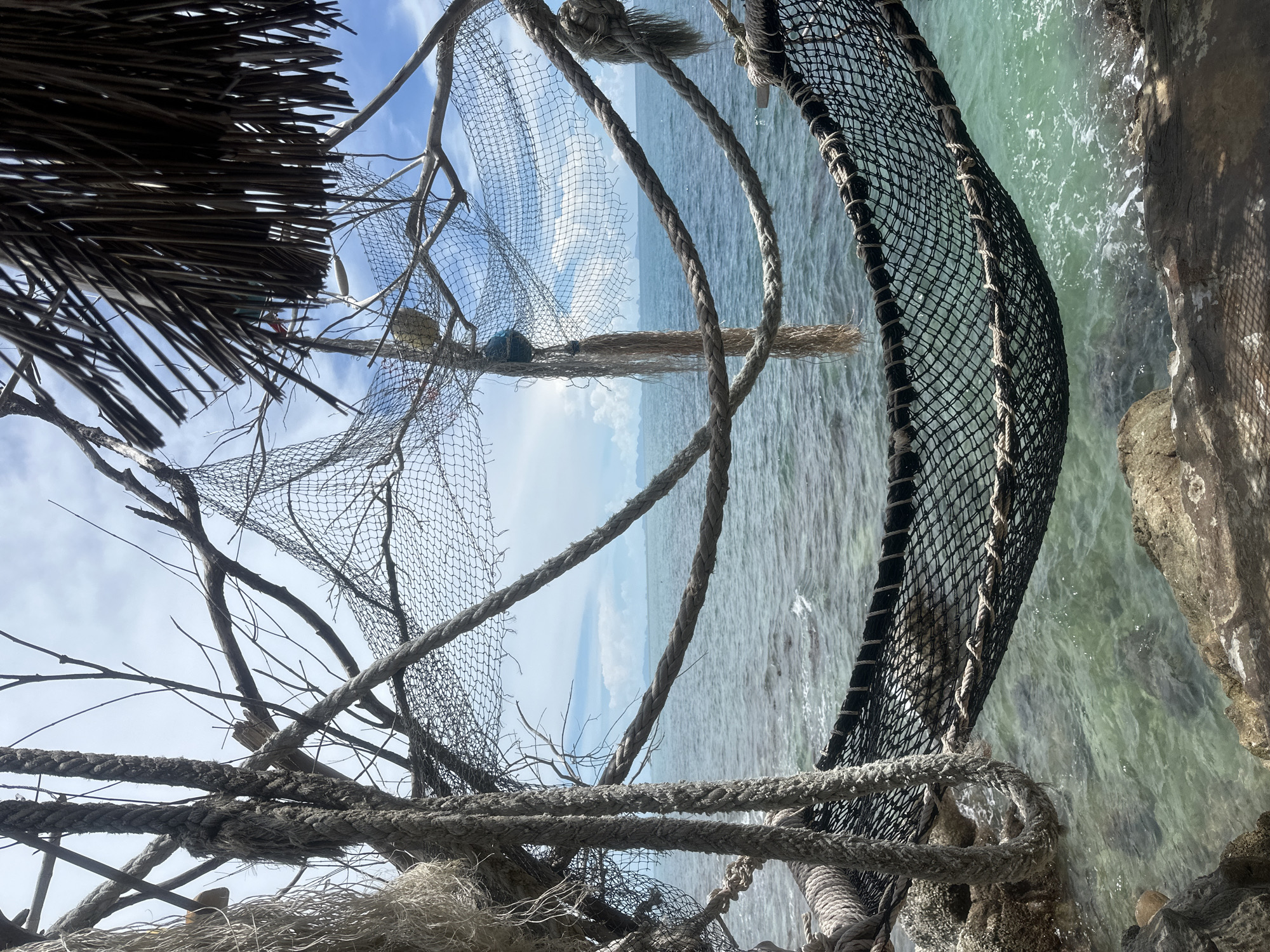 This media file depicts a close-up, angled view of fishing nets and ropes in the foreground, with a body of water and sky in the background. The scene appears to be outdoors, likely near a coast or on a boat. The nets are made of mesh and are supported by branches and thick ropes. Some of the nets are tangled, suggesting they are either in use or have been discarded.
The water is a clear, greenish-blue, with some ripples and a rocky shoreline visible on the right side of the frame. The sky is bright blue with scattered white clouds, indicating a sunny day. The lighting suggests it is daytime, possibly late morning or early afternoon.
The overall impression is one of a rugged, maritime environment, possibly a fishing village or a secluded beach area. The materials—nets, ropes, branches, and rough-hewn wood—create a rustic and weathered aesthetic. There are no people visible in the media file.
The focus is on the textures and materials of the fishing gear, contrasting with the natural elements of the water and sky. The composition is dynamic due to the angled perspective and the intersecting lines of the nets and ropes. The location context of Koh Phangan, Thailand, evokes a tropical coastal setting, which aligns with the visual cues in the image. There is no visible text in the media file.